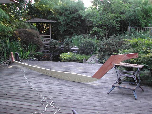 Partially assembled wooden boat frame on a deck, supported by a workbench, with a pond and garden in the background.
