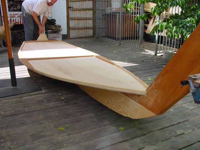A person is assembling a wooden boat on a deck, adjusting a large panel into place.