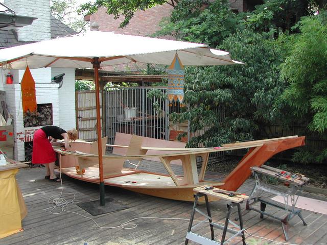 A person is working on a partially built wooden boat under a large umbrella in an outdoor workspace.