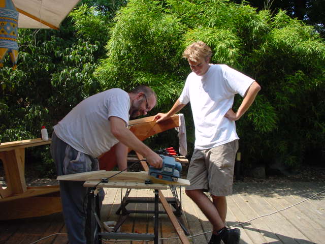 One person uses a power saw to cut wood while another watches, both working on a boat project outdoors.