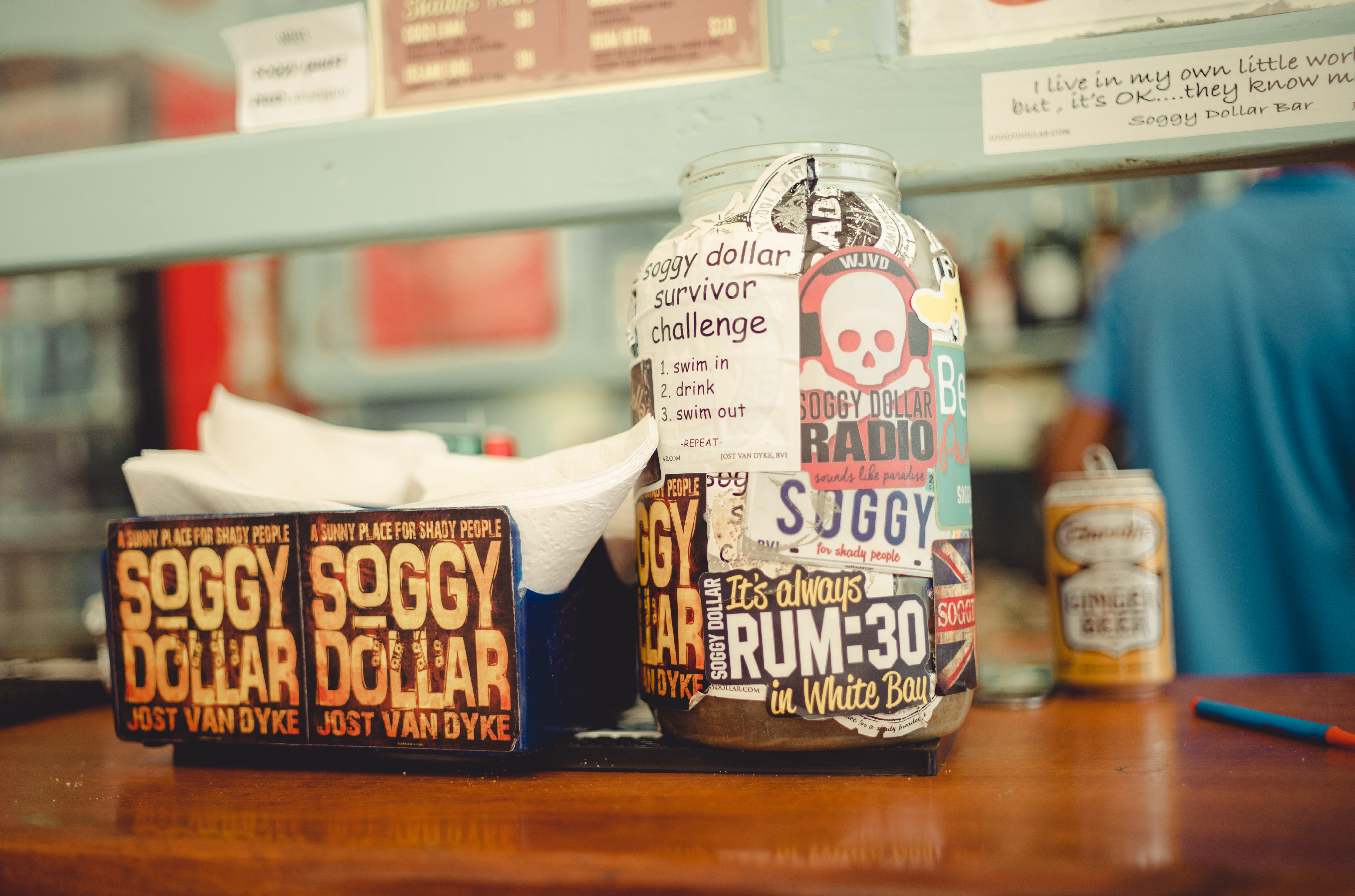 A glass jar covered in stickers promotes the "Soggy Dollar Survivor Challenge" on a bar counter with napkins.