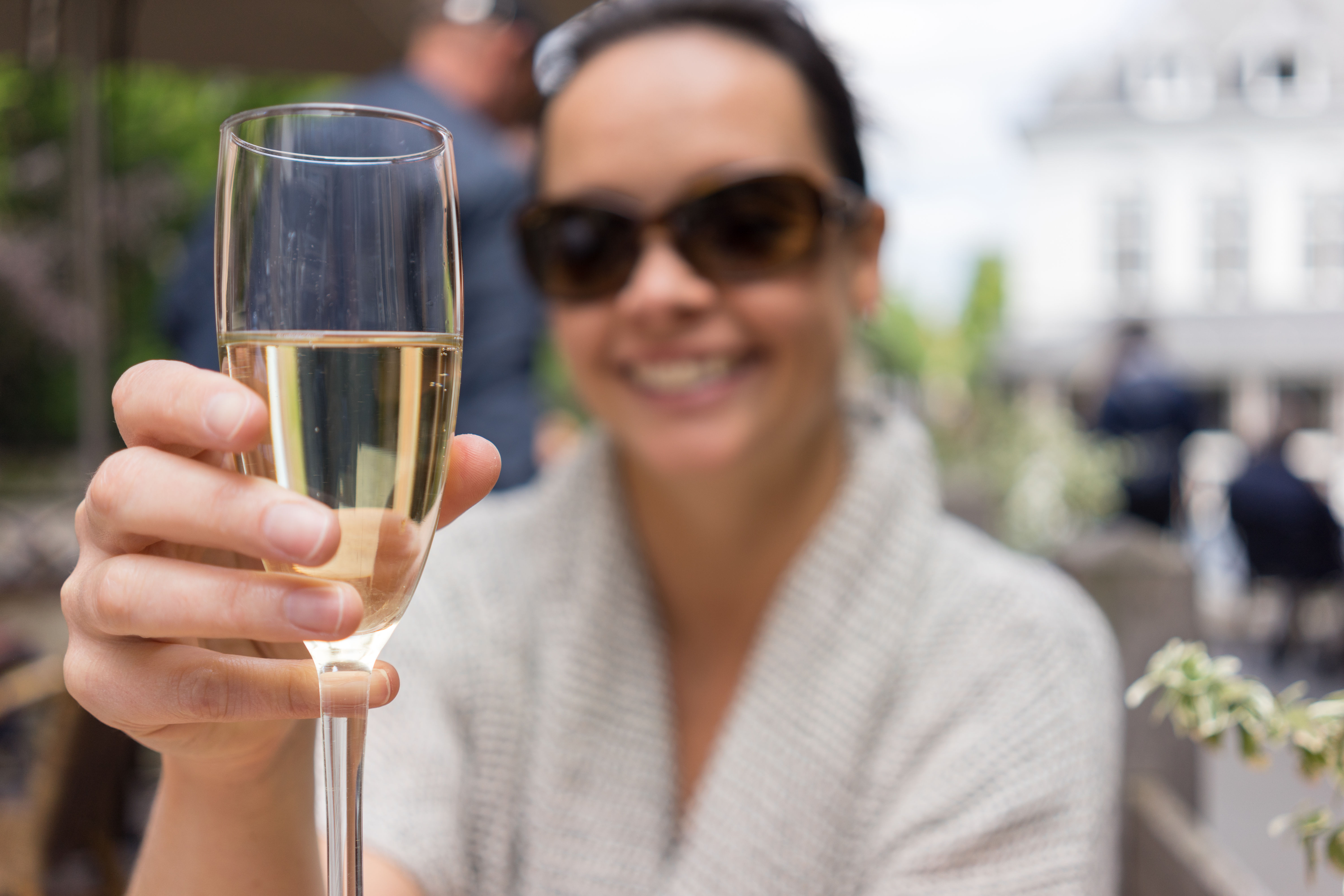 A person wearing sunglasses smiles while holding up a glass of sparkling wine in an outdoor setting.