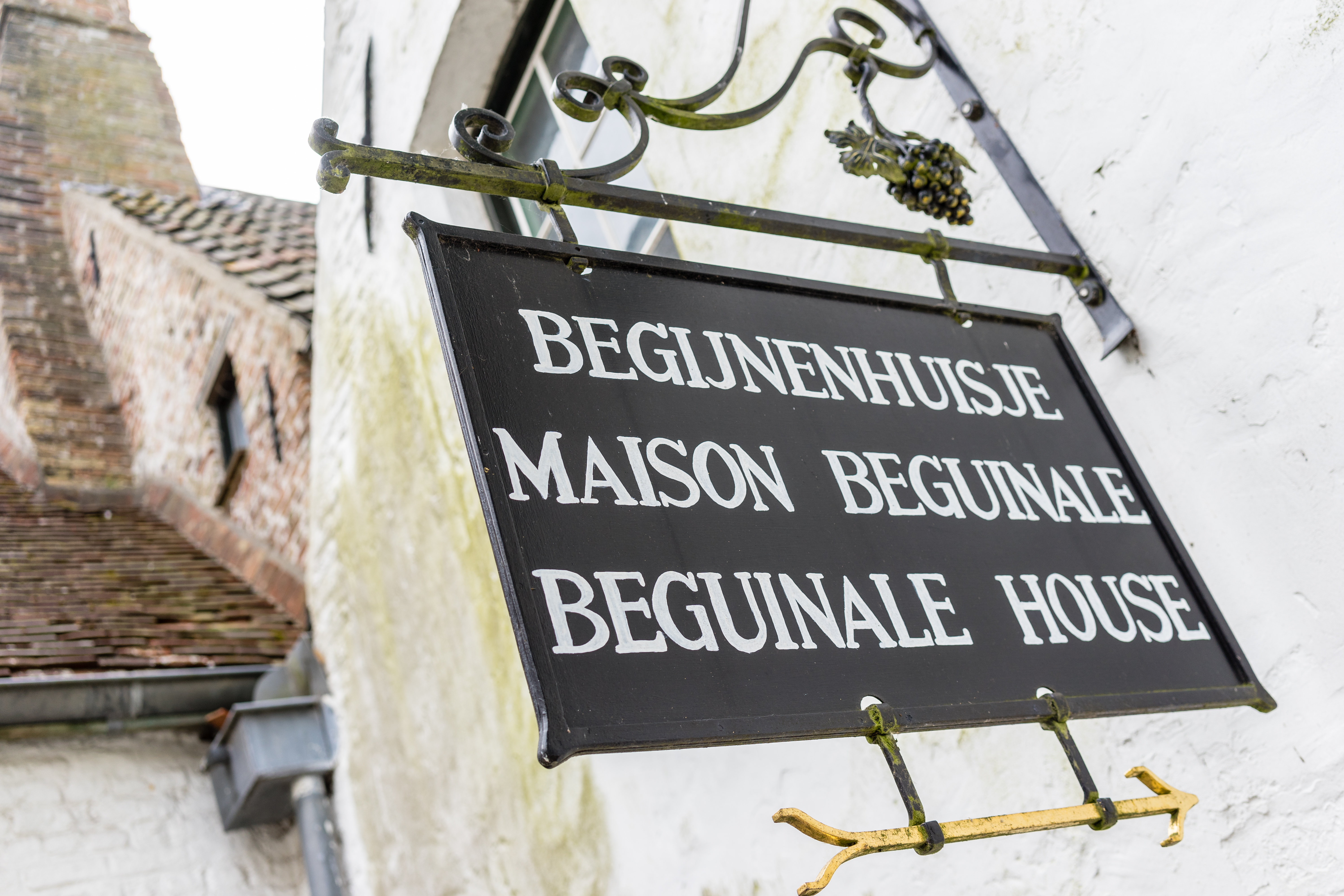 A black metal sign with white lettering reads "Beguinale House" in three languages, hanging outside a historic building.
