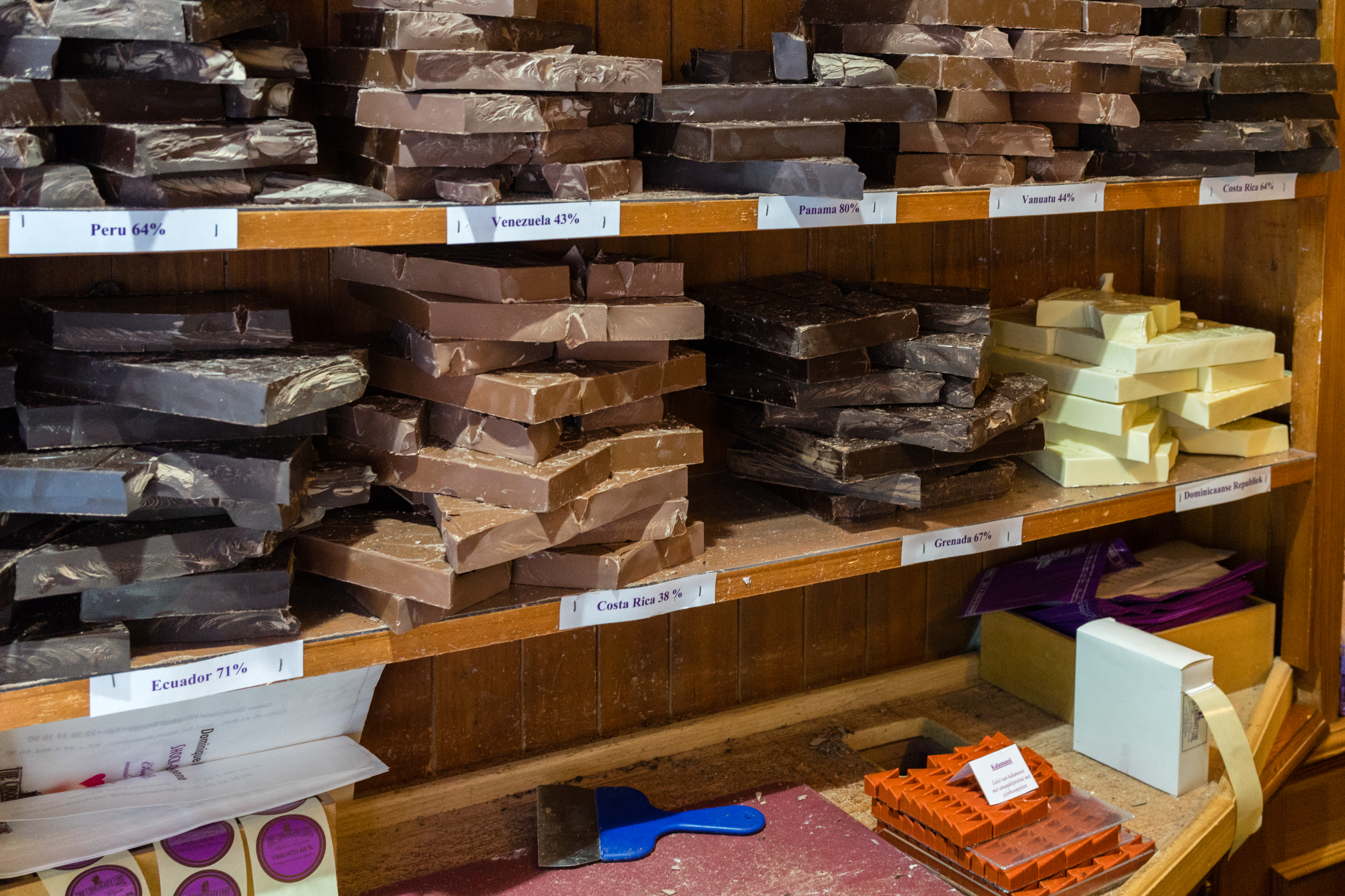 Stacks of dark, milk, and white chocolate bars labeled by cocoa origin on wooden shelves in a chocolate shop.