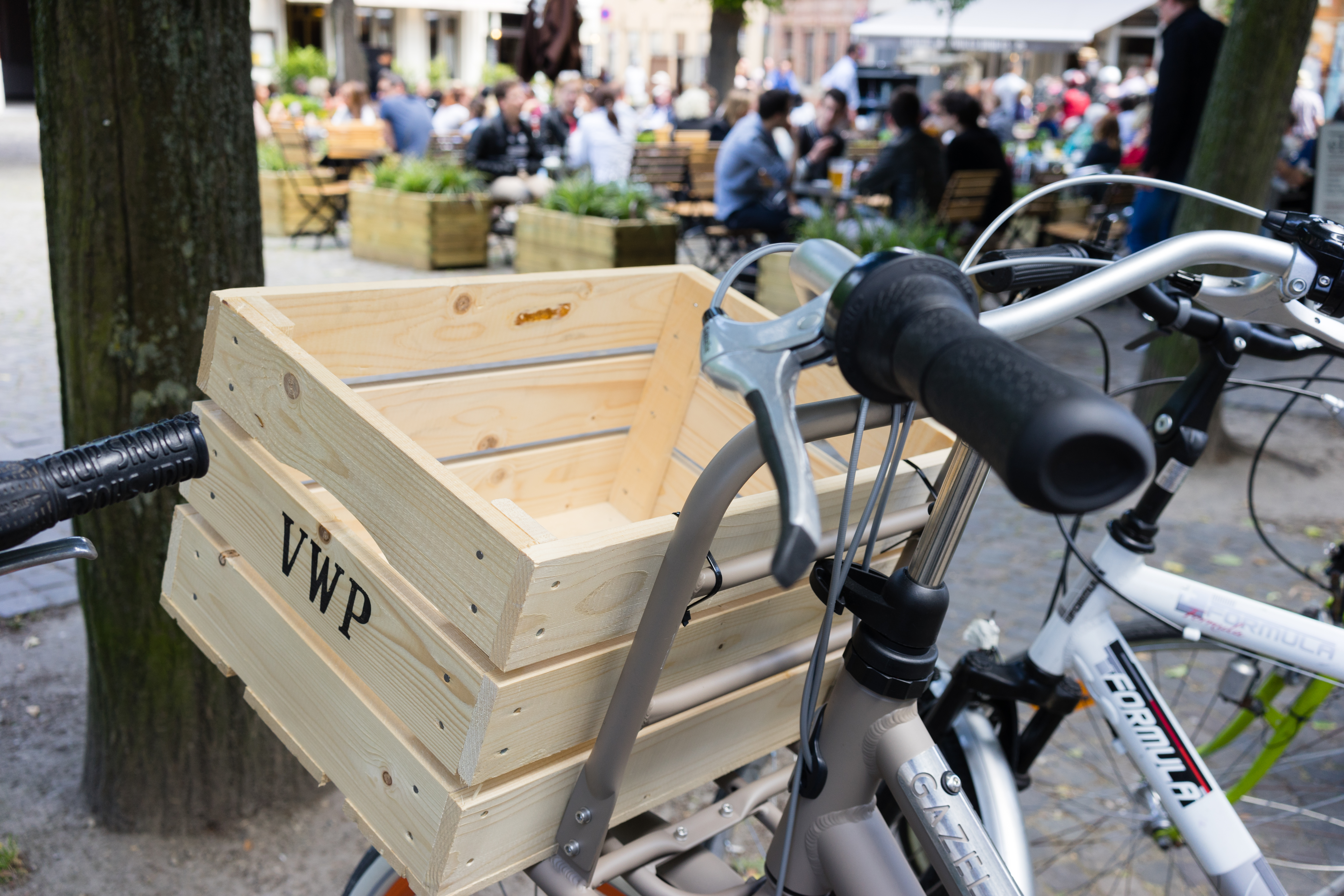 A bicycle with a wooden crate attached to the front, parked near an outdoor café with people in the background.