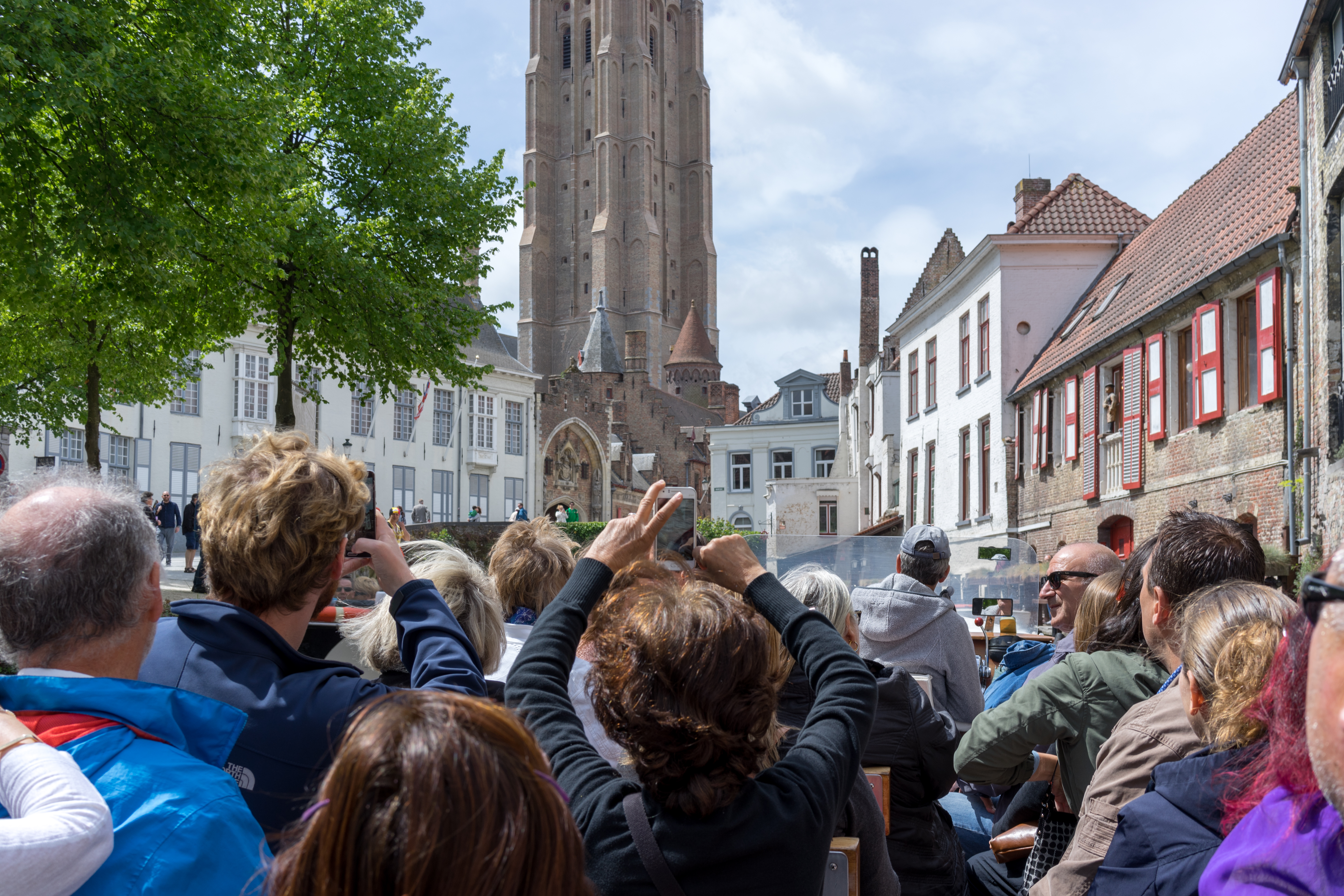 A group of tourists on a boat tour in Bruges, taking photos of a tall historic church.