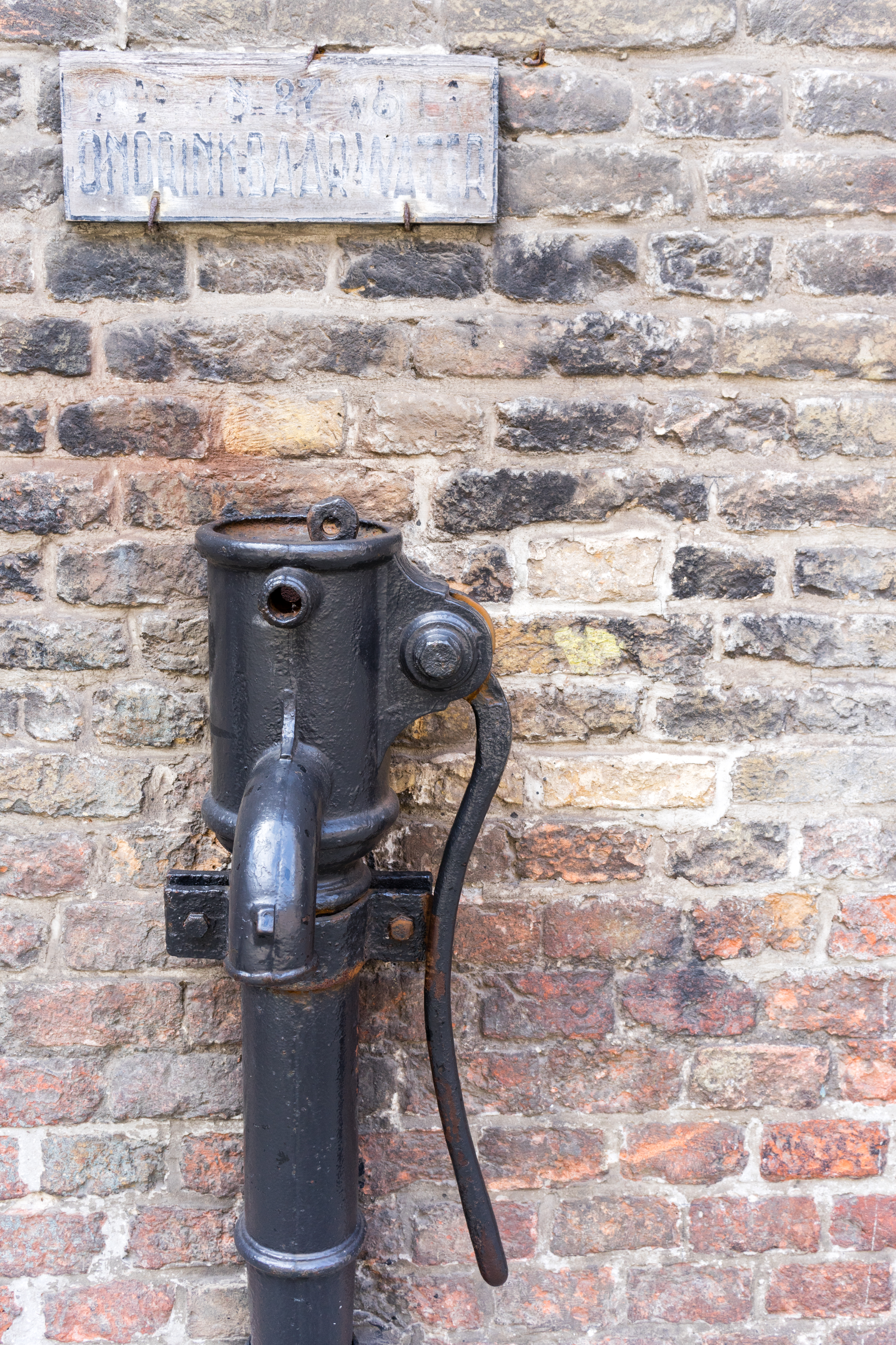 An old black metal water pump with a long handle, mounted on a brick wall with a worn sign above.