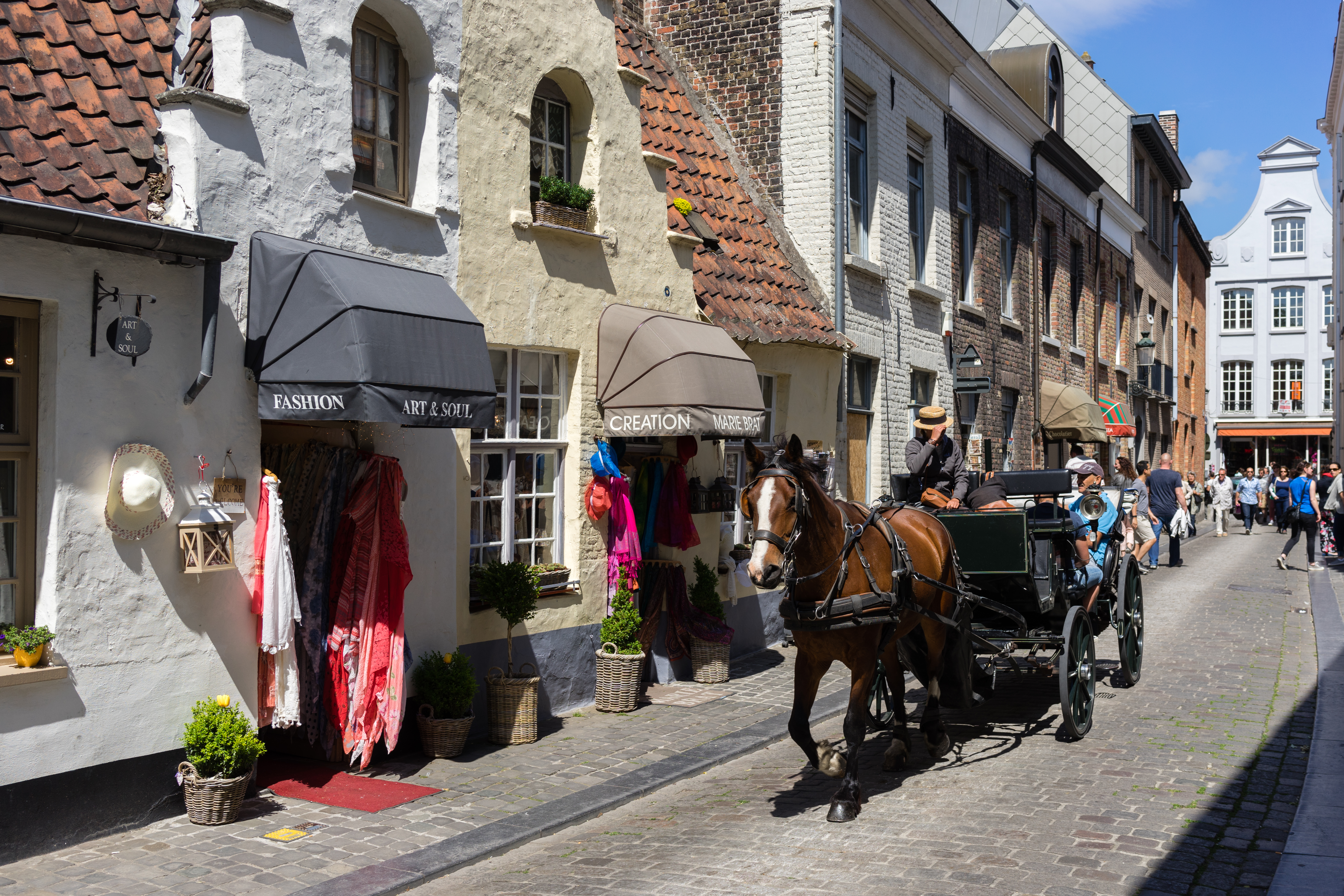 A horse-drawn carriage passes by small shops with colorful clothing displays on a busy shopping street in Bruges.