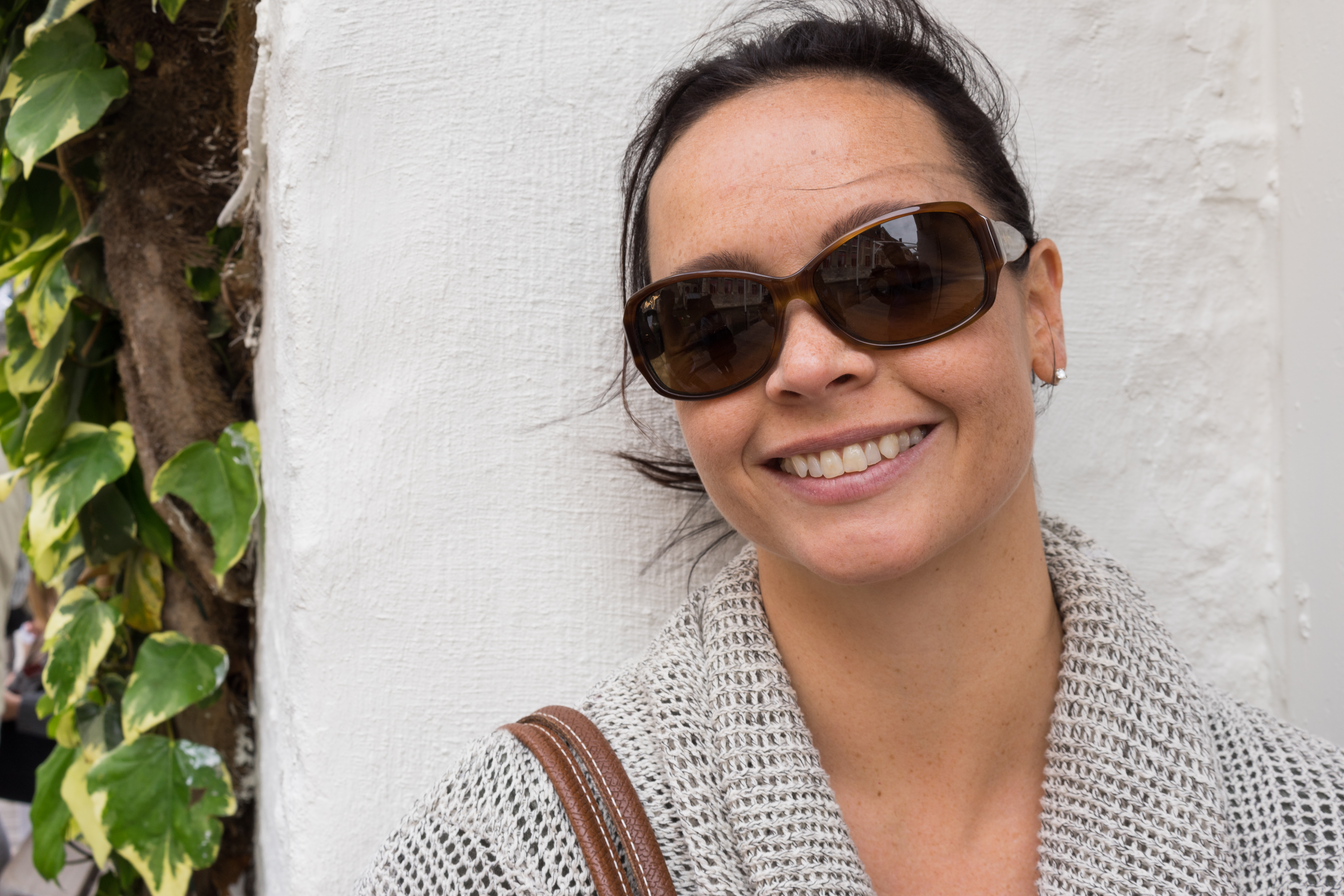 A woman wearing sunglasses and a knitted sweater smiles while leaning against a white wall.