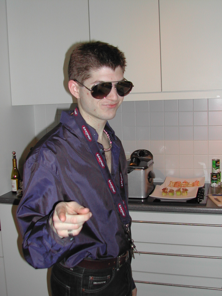 A person wearing sunglasses and a shiny shirt points at the camera while standing in a kitchen.