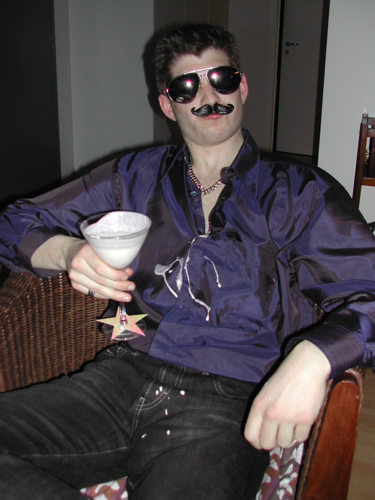 A person in a costume with sunglasses and a fake mustache holds a cocktail, with spilled drink on their shirt.
