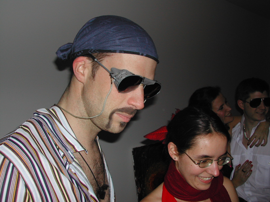 A man in a striped shirt and bandana wears unique sunglasses, while others in costumes smile and talk nearby.