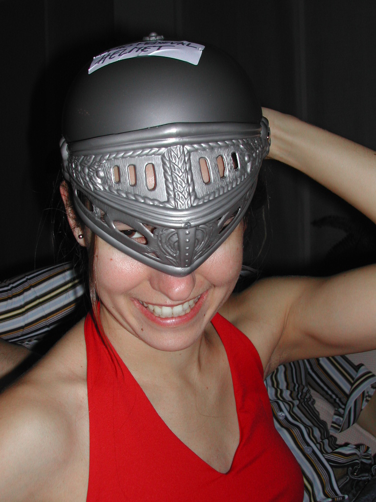 A smiling person in a red top wears a plastic knight helmet tilted over their face at a carnival event.