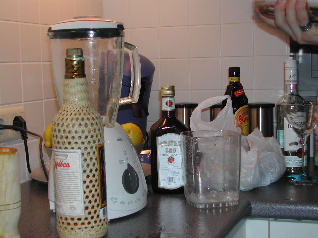 A kitchen counter with a blender, bottles of alcohol, a glass with ice, and a hand pouring a drink.
