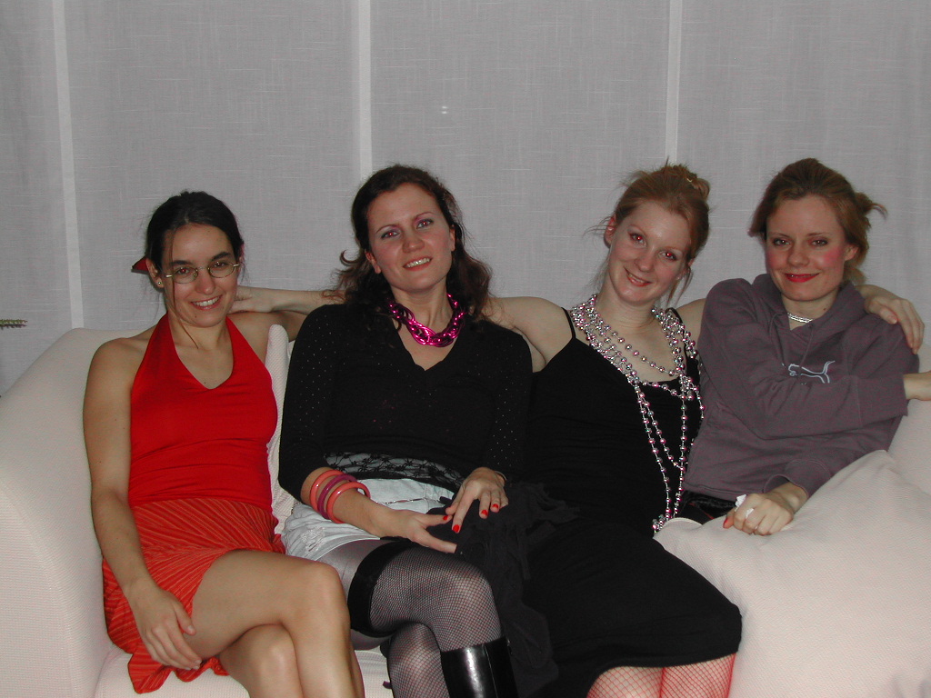 Four women in festive outfits sit closely on a white couch, smiling at the camera during a Carnaval celebration.