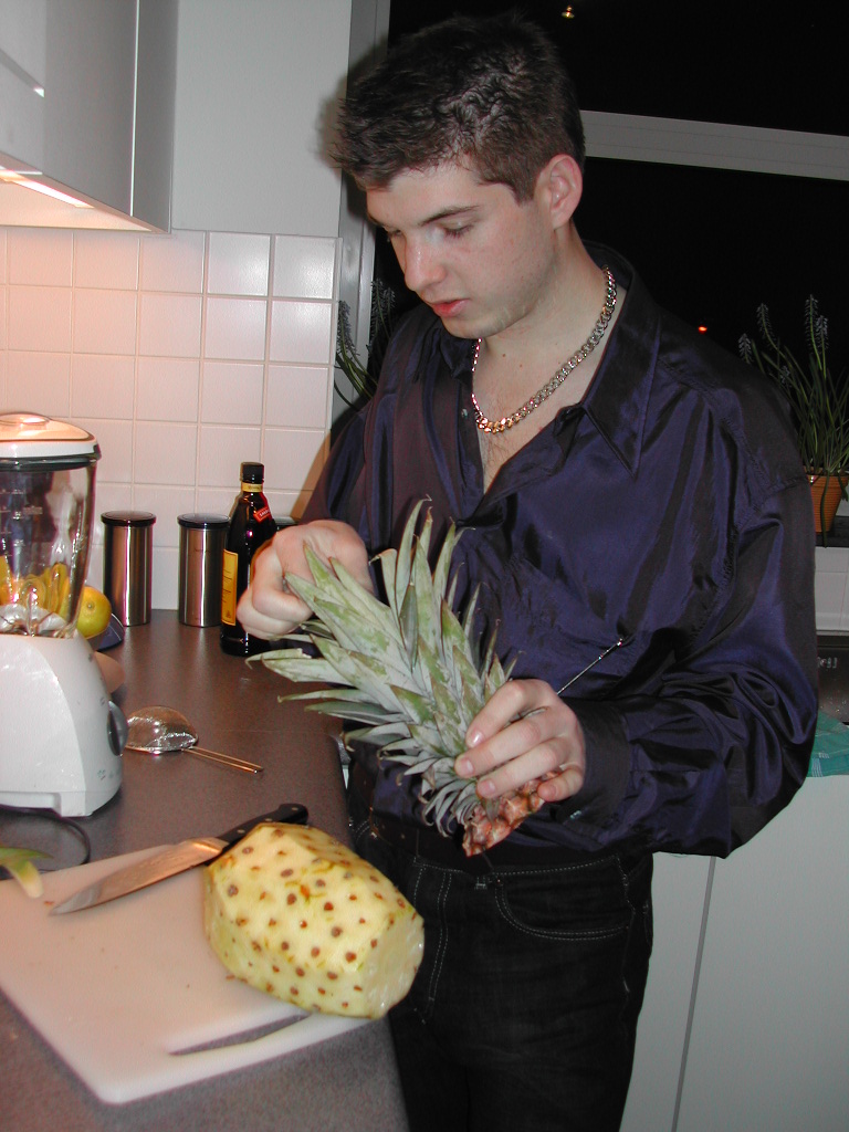 A man in a shiny dark shirt prepares a pineapple in a kitchen, holding the top while cutting.