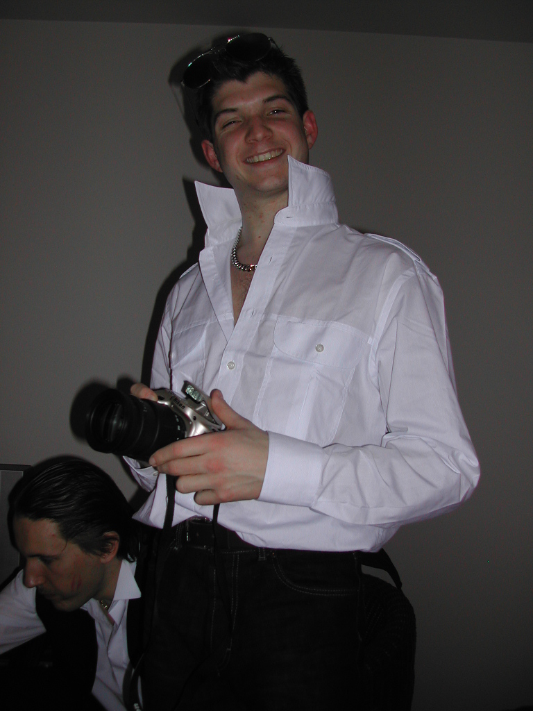 A smiling person in a white shirt holds a camera, while another person sits in the background.