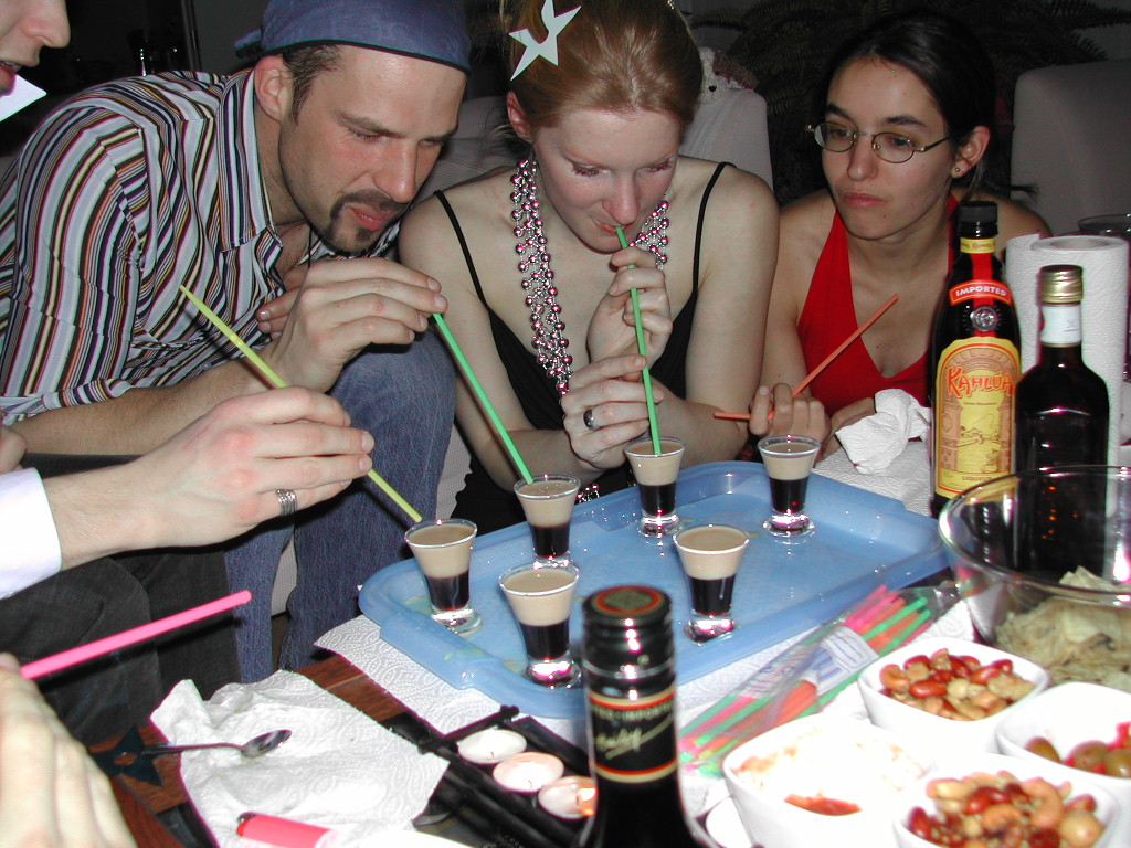 A group of people use straws to drink layered shots from a tray at a party.