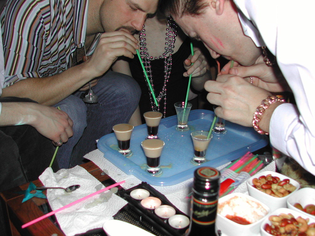 A group of people drinks shots through straws from a tray at a party, surrounded by snacks and drinks.