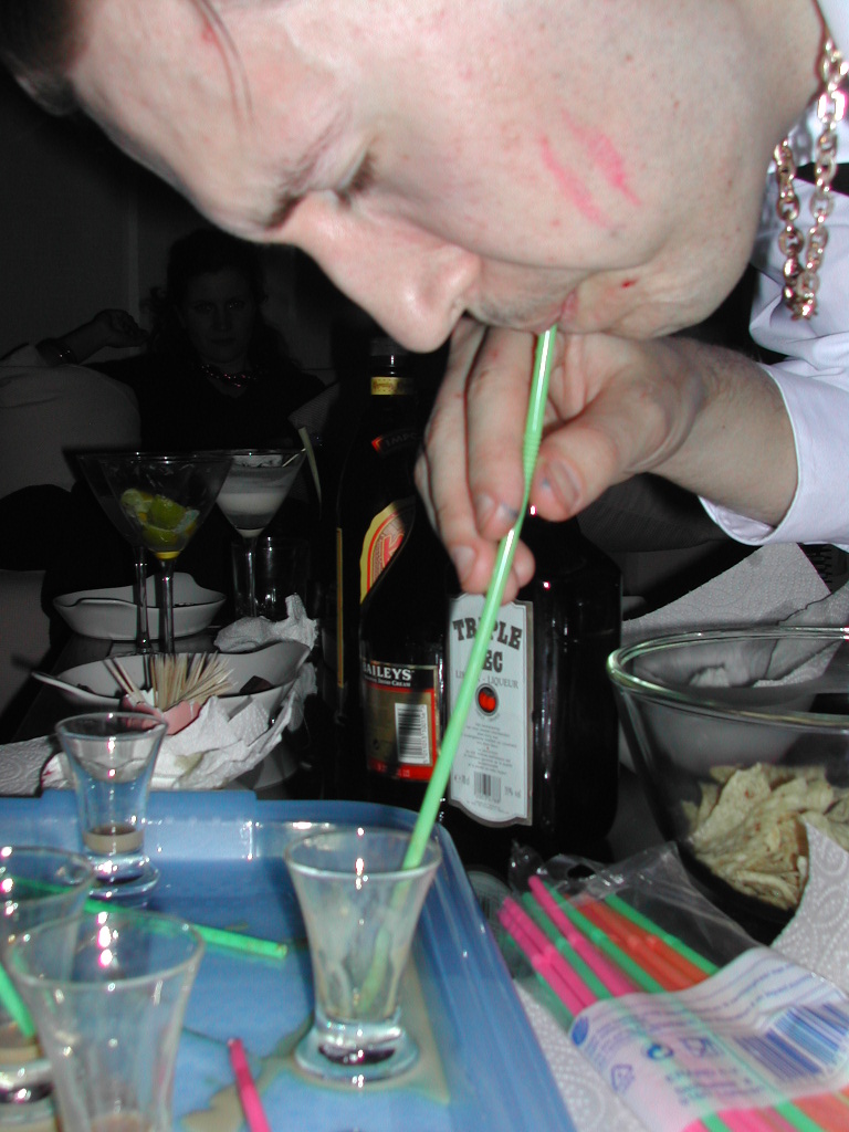 A man leans over a tray, drinking from a shot glass with a straw at a party.