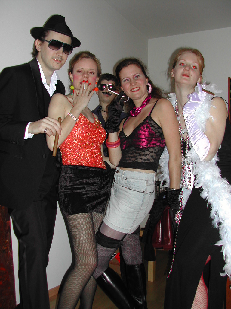 A group of five people in festive costumes posing together, some holding cigars and wearing accessories.