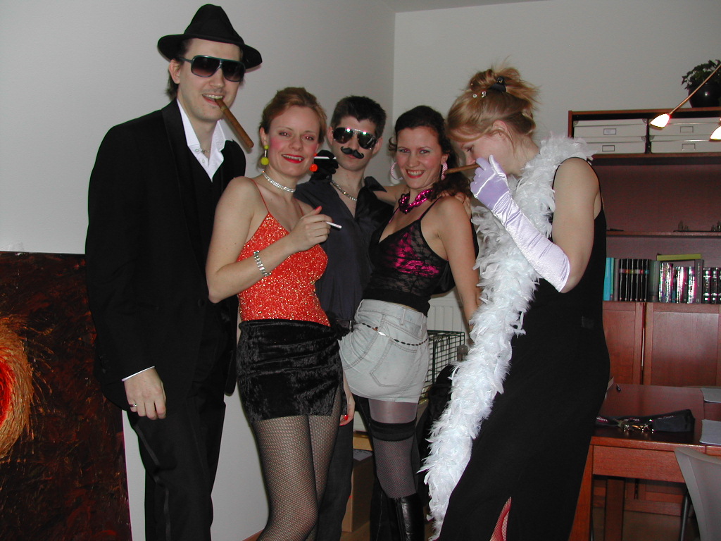 Five people in festive costumes pose together indoors, some holding cigars and cigarettes, dressed for a carnival celebration.