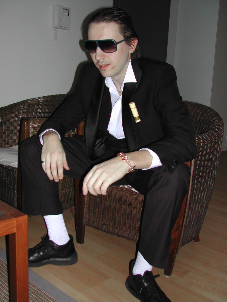 A person in a black suit and sunglasses sits on a chair, holding a relaxed posture with a prop cigar.