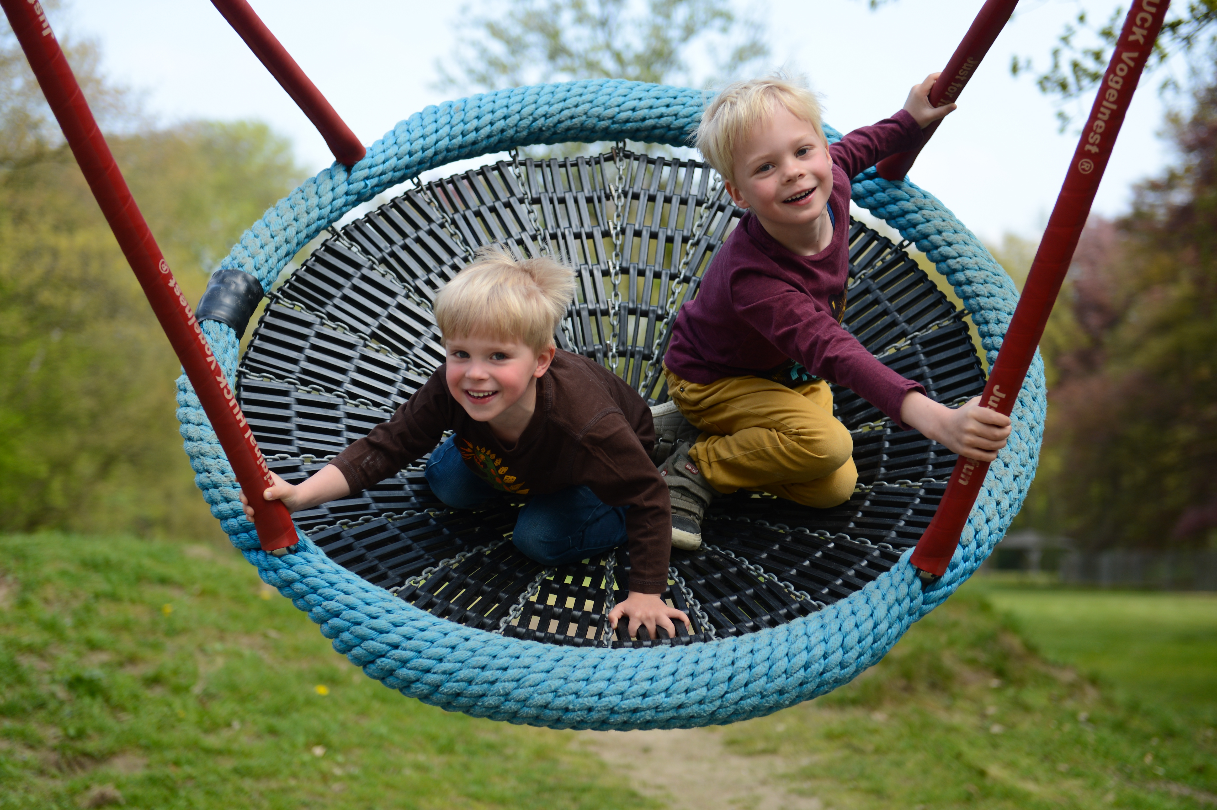 Two young children smile while sitting on a large rope swing at a playground.