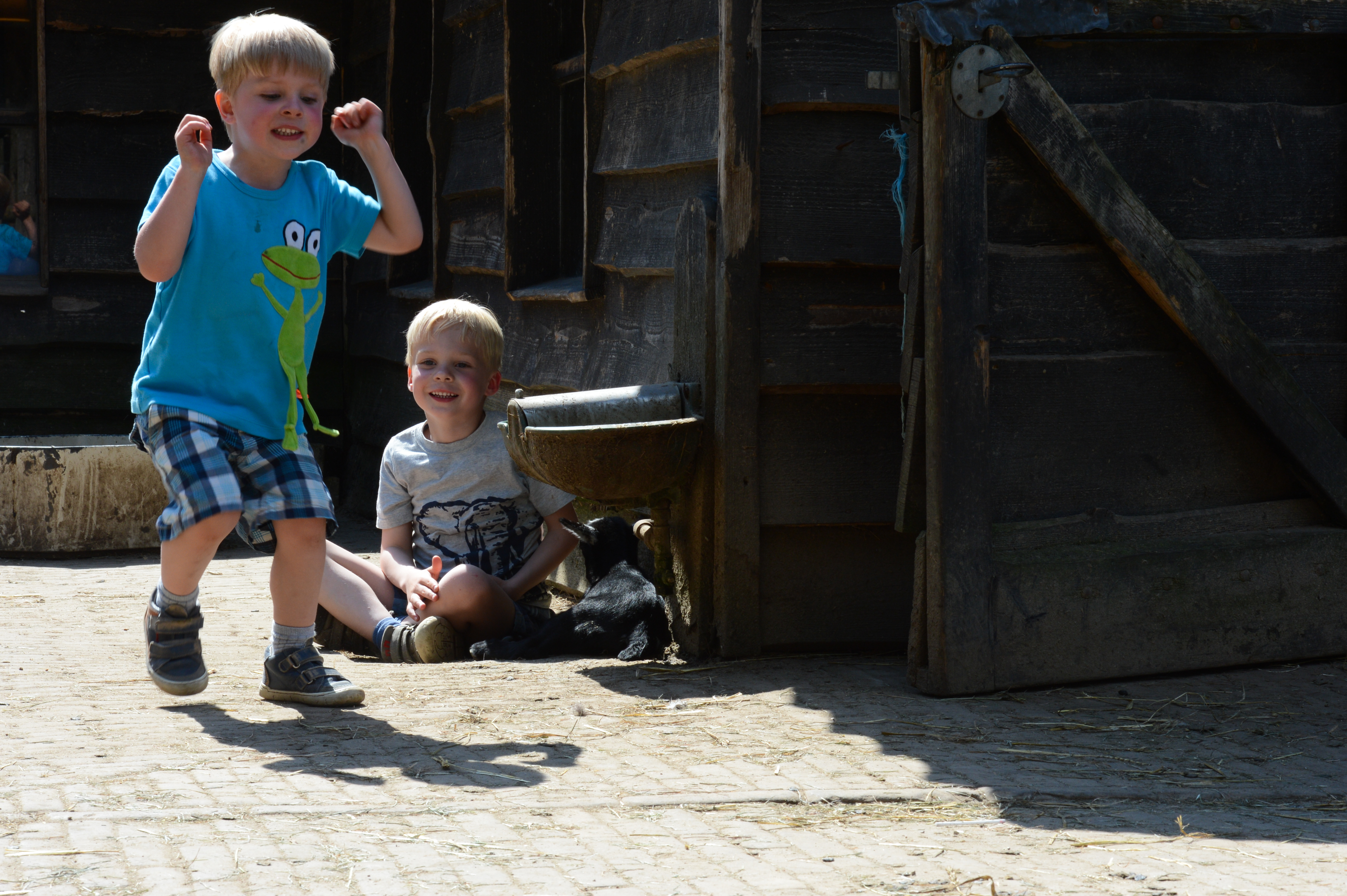 A young boy jumps with excitement while another boy sits on the ground, smiling near a wooden barn.