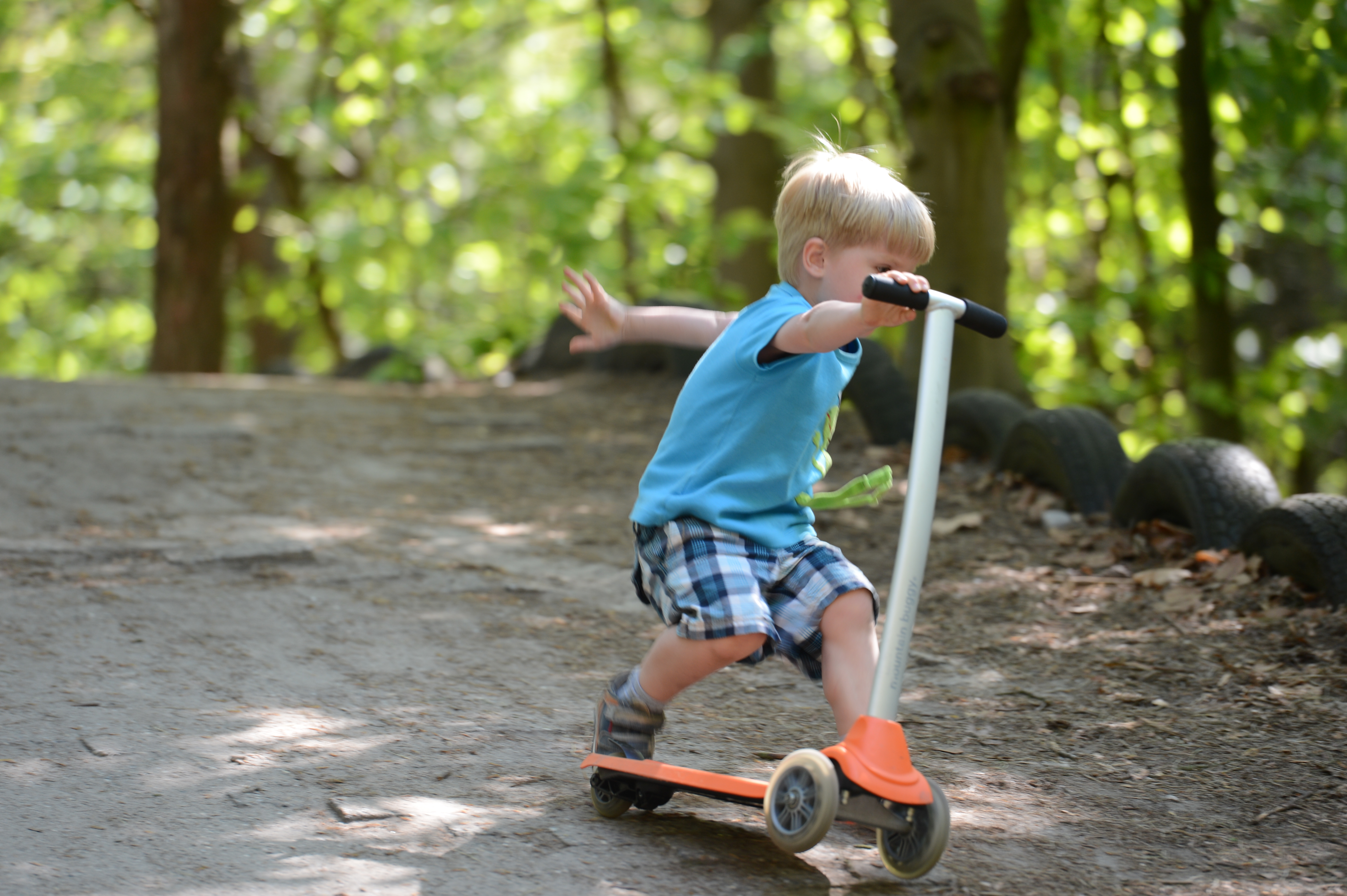 A young child in a blue shirt rides a scooter on a dirt path, balancing with one foot.
