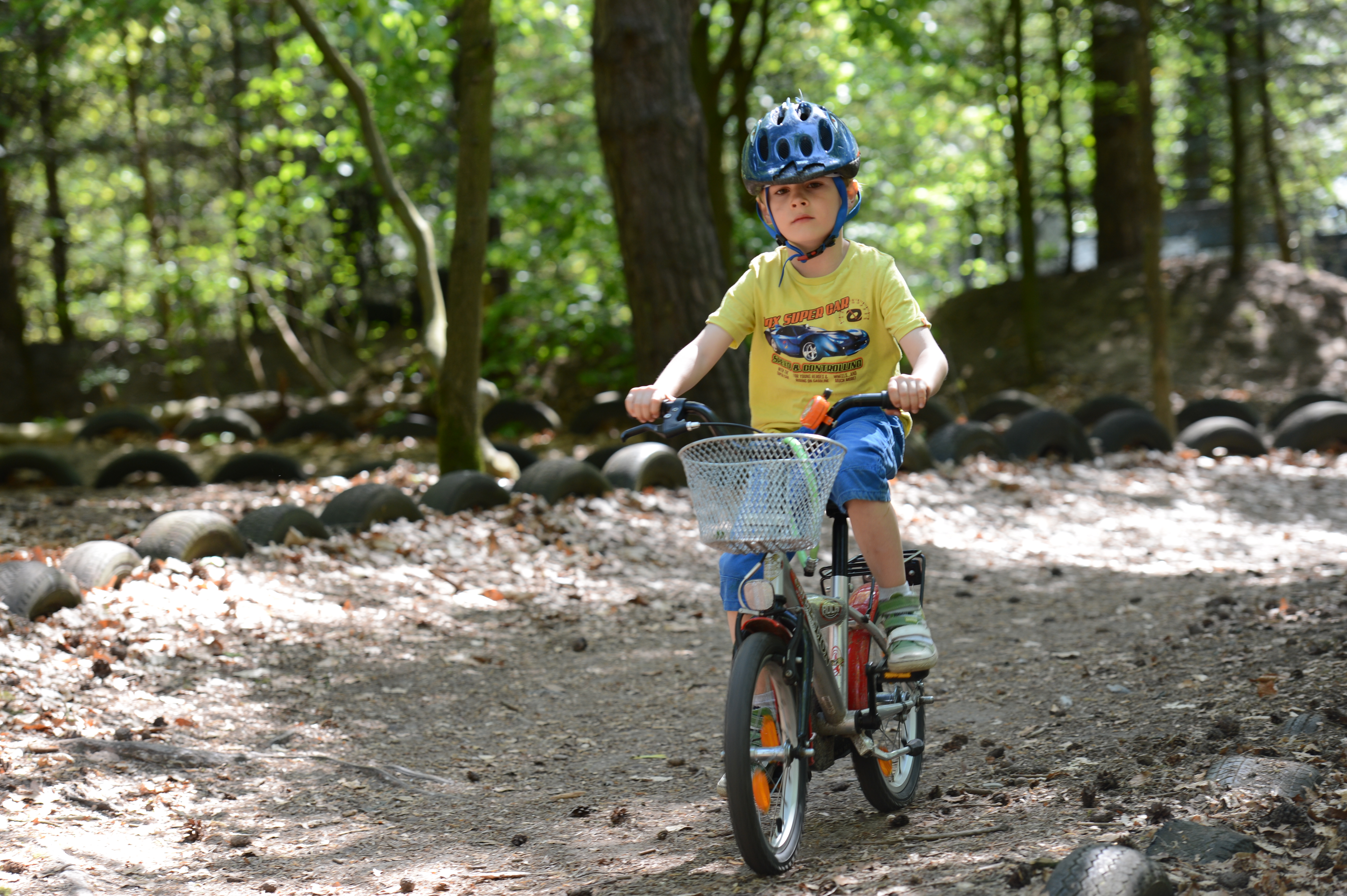 A young child wearing a helmet rides a bicycle on a dirt path surrounded by trees and tires.