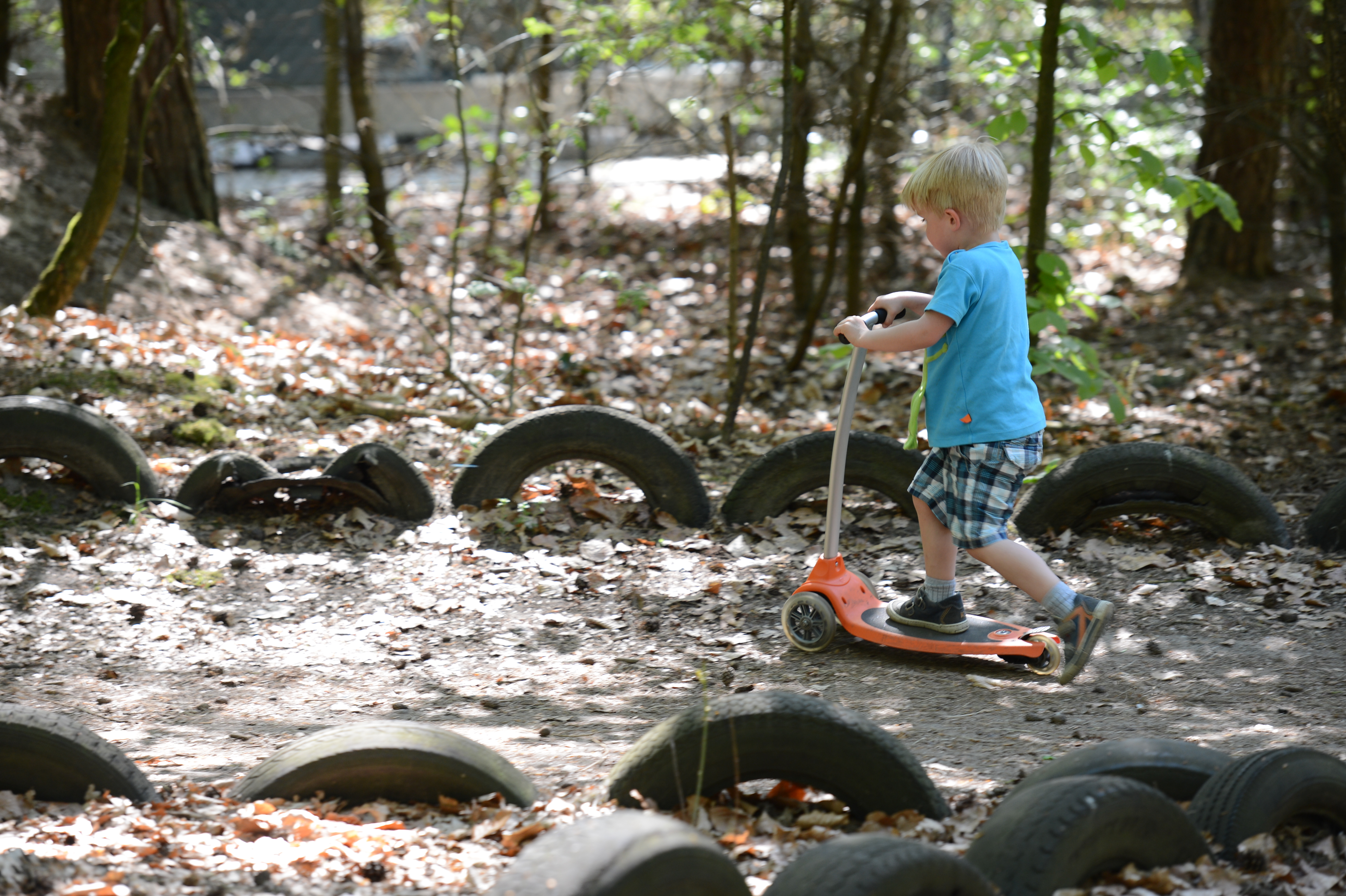 A young child rides an orange scooter on a dirt path surrounded by half-buried tires in a wooded area.