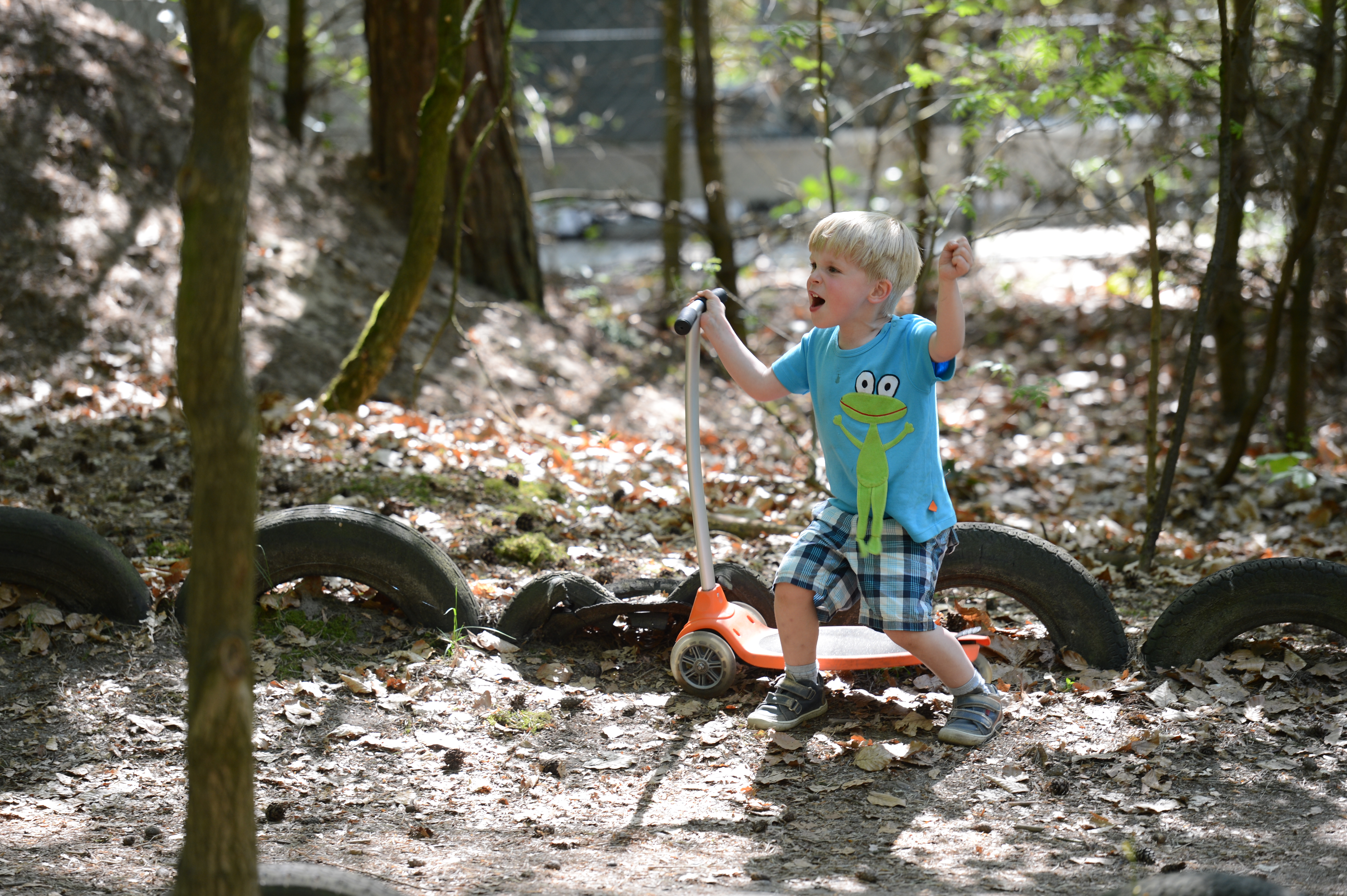 A young child in a blue frog shirt rides an orange scooter on a dirt path surrounded by trees and tires.