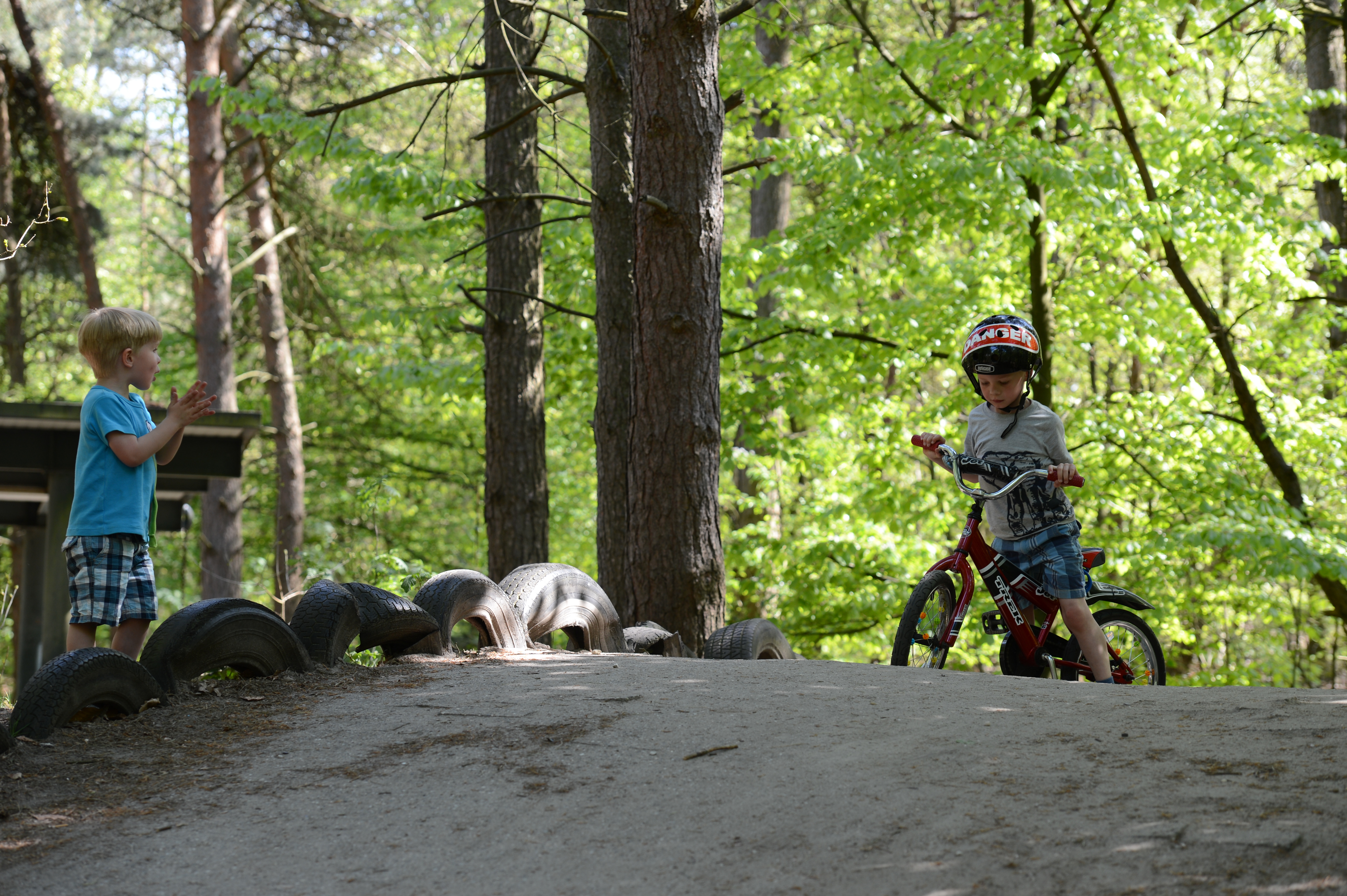 A child wearing a helmet rides a bicycle down a dirt path while another child claps nearby.