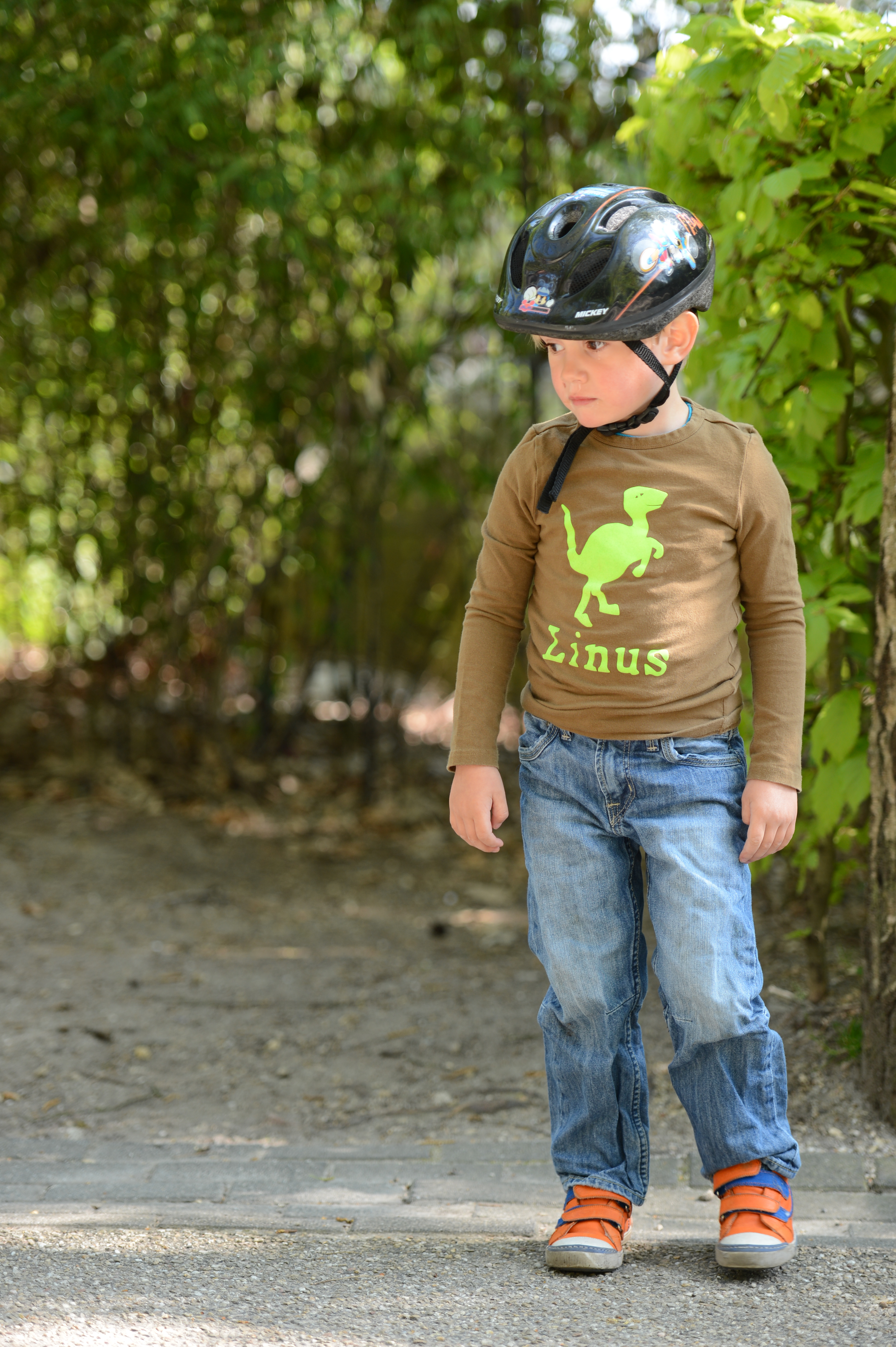 A young child wearing a bike helmet and casual clothes stands on a path, looking to the side.