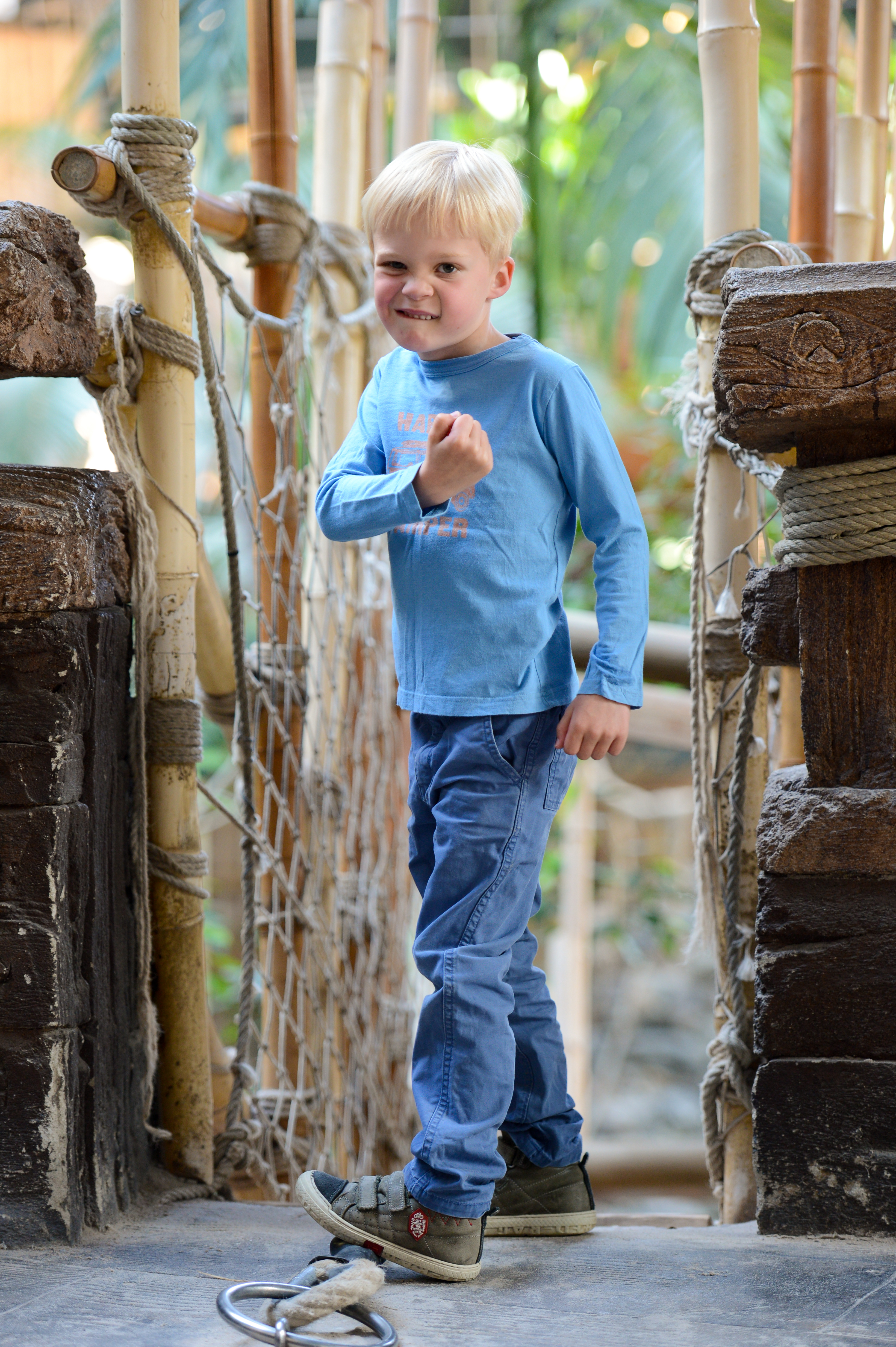 A young child in a blue shirt stands on a wooden bridge, smiling and making a playful fist gesture.