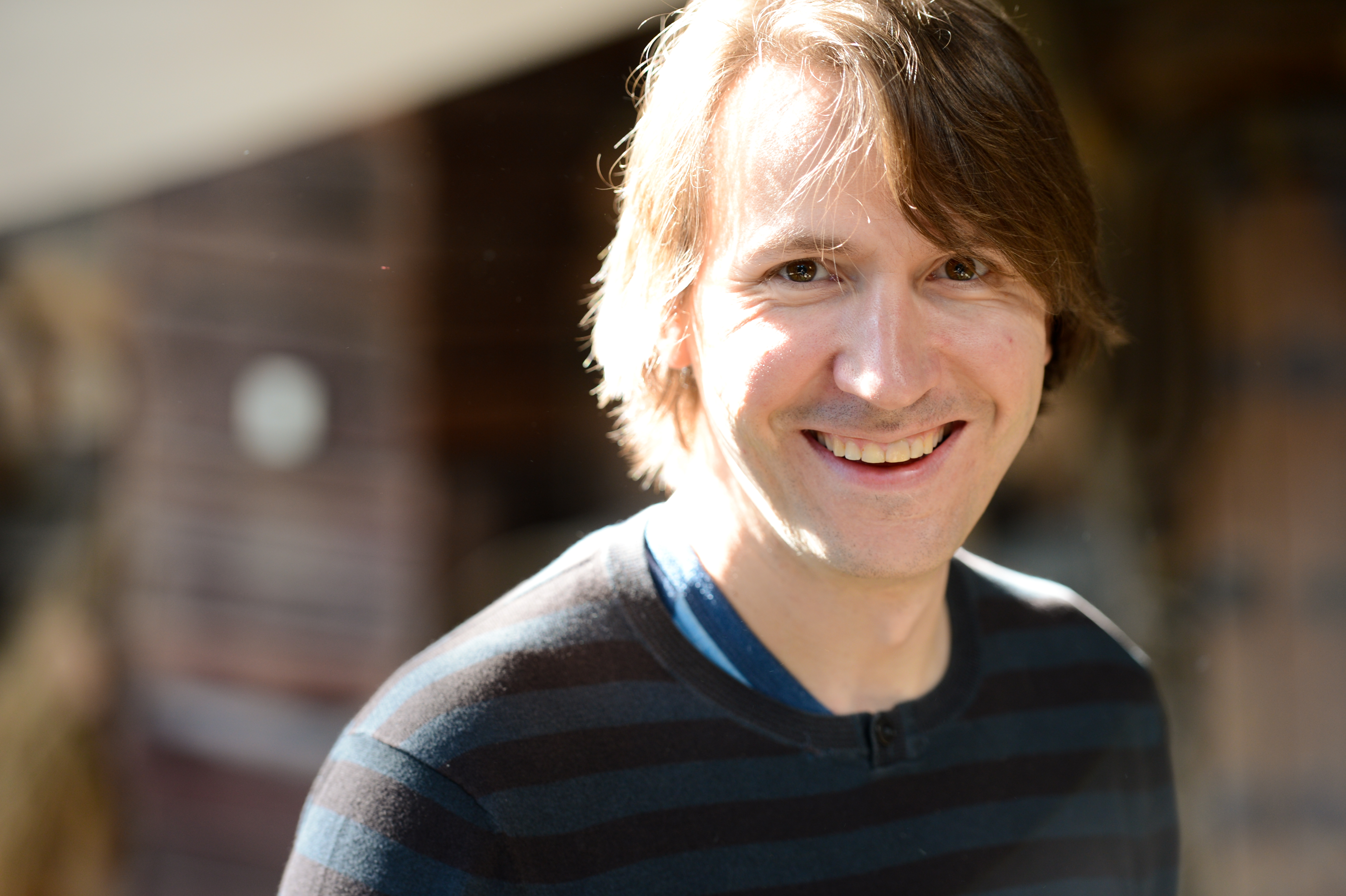A person with medium-length hair and a striped sweater smiles at the camera in natural lighting.