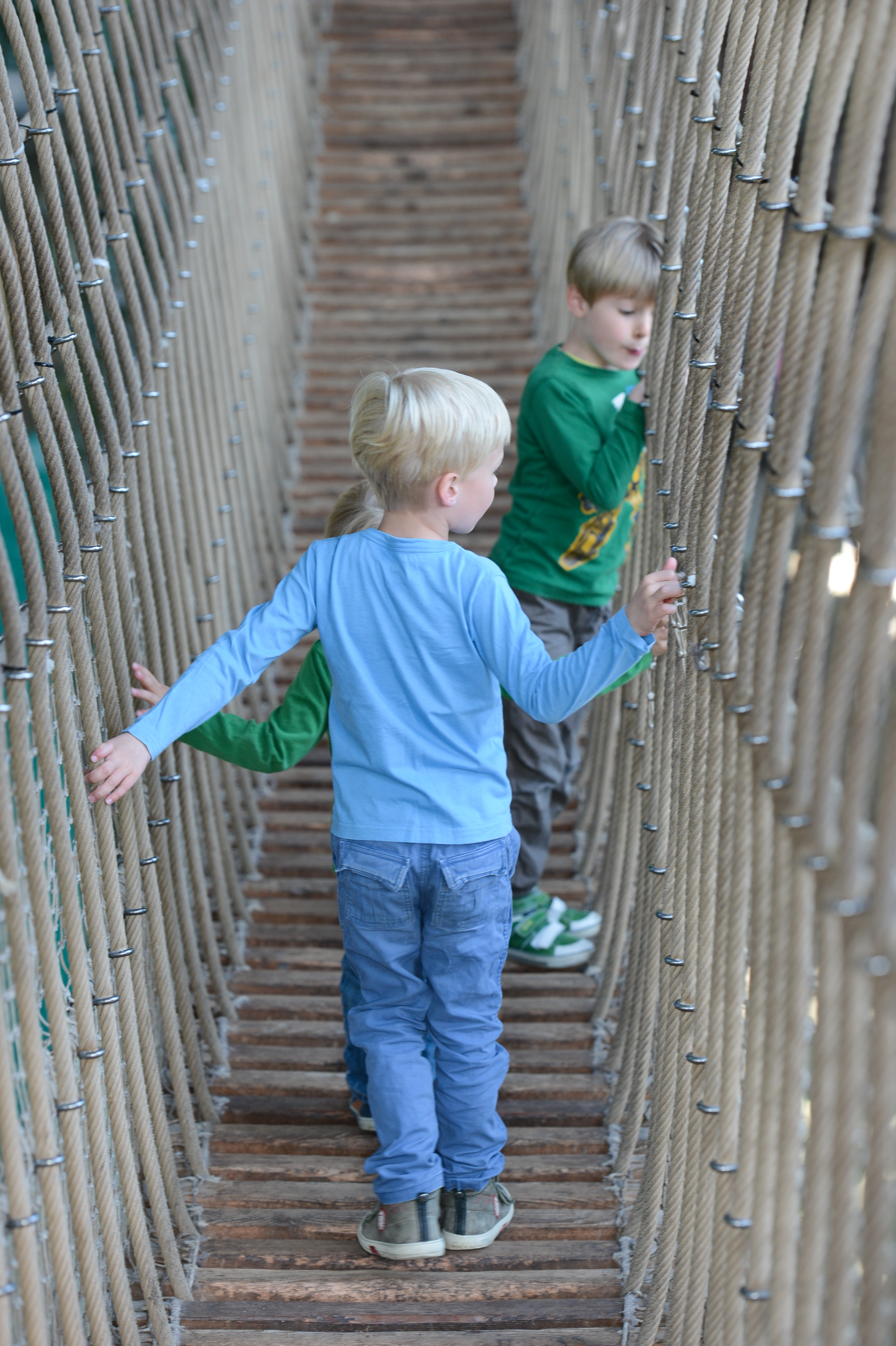 Three children walk across a rope bridge, holding onto the sides for balance.