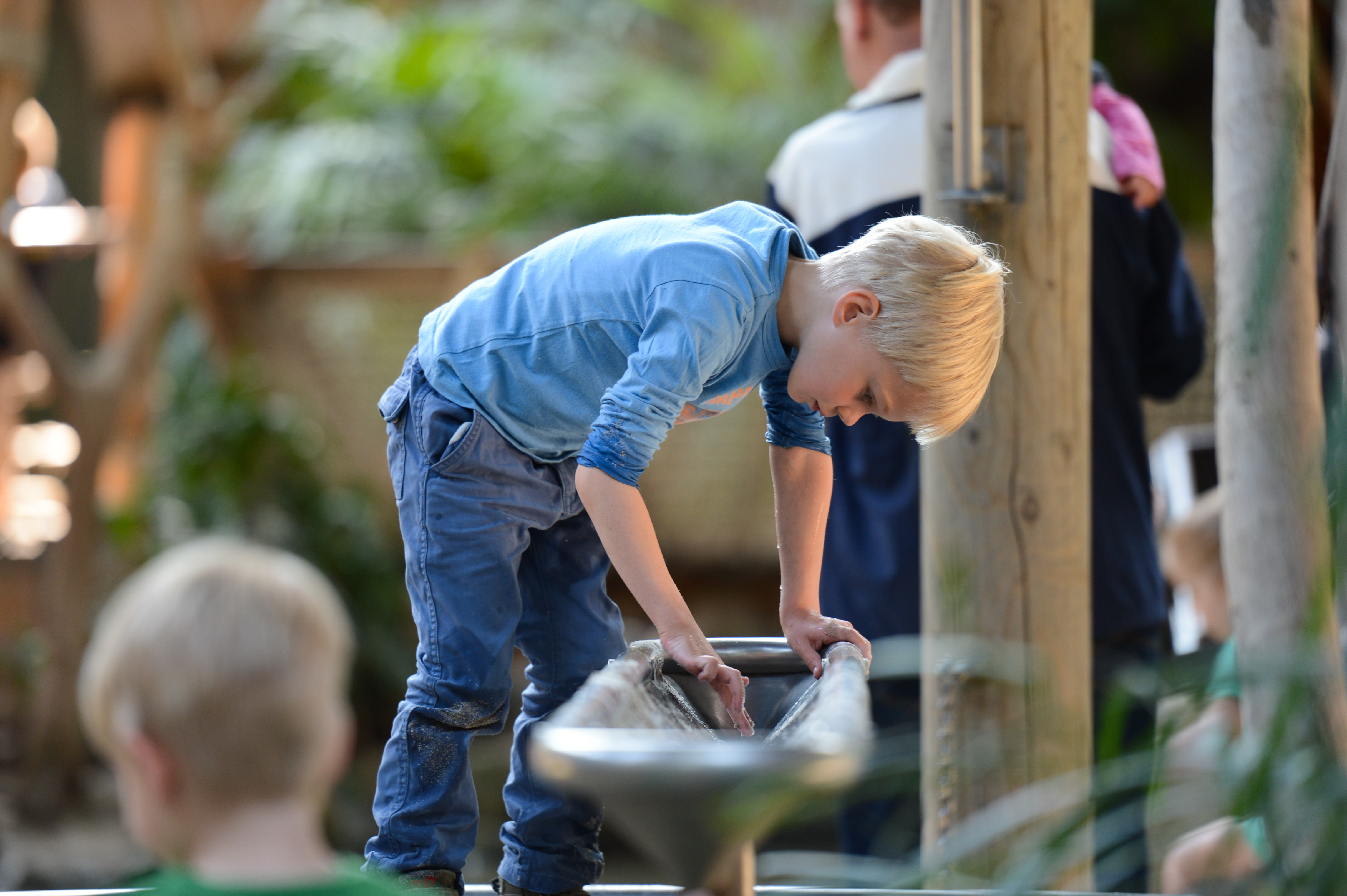 A young boy in a blue shirt leans over a water play structure, interacting with the flowing water.
