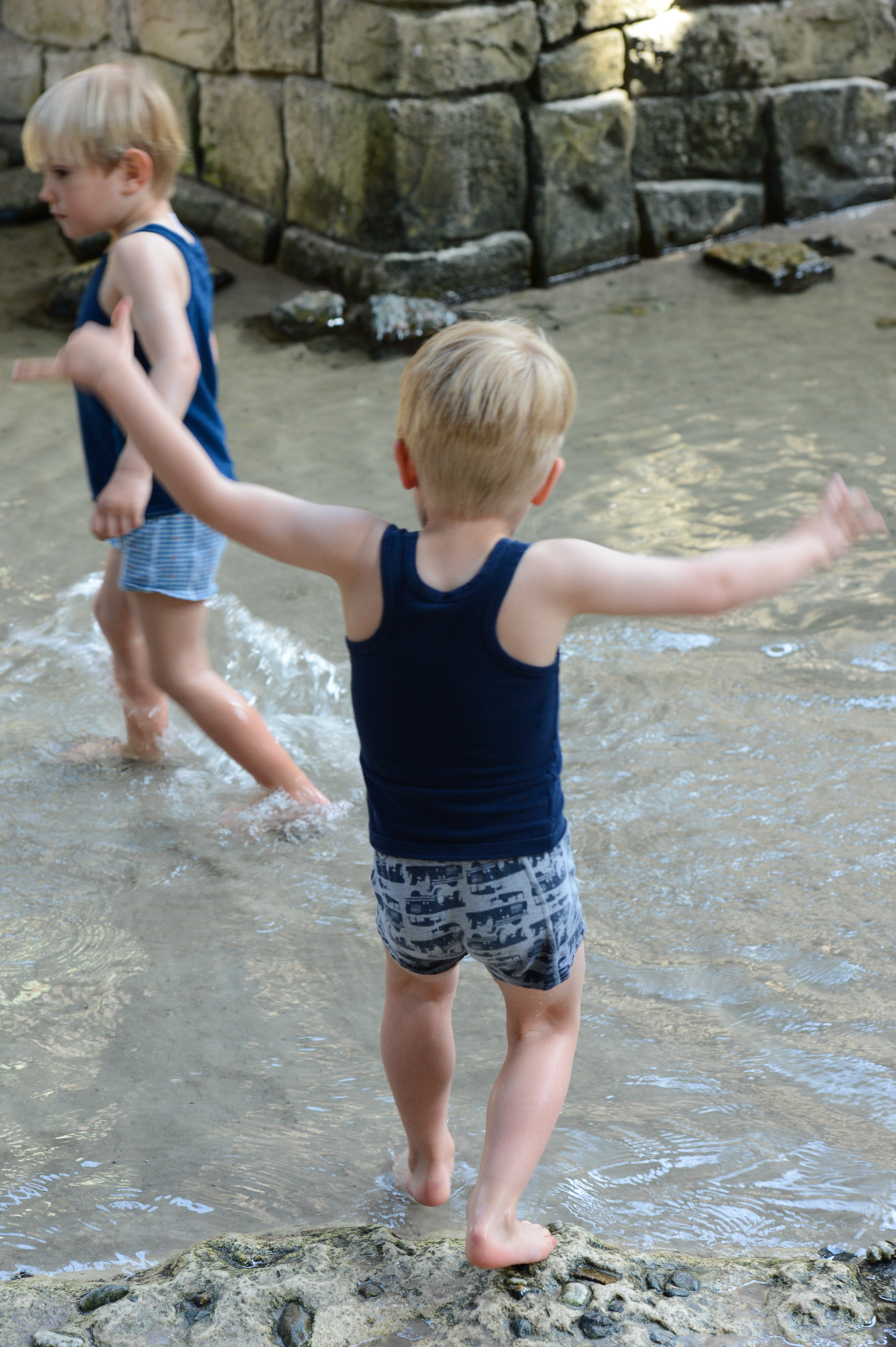 Two young children wade through shallow water, splashing and playing near a stone wall.