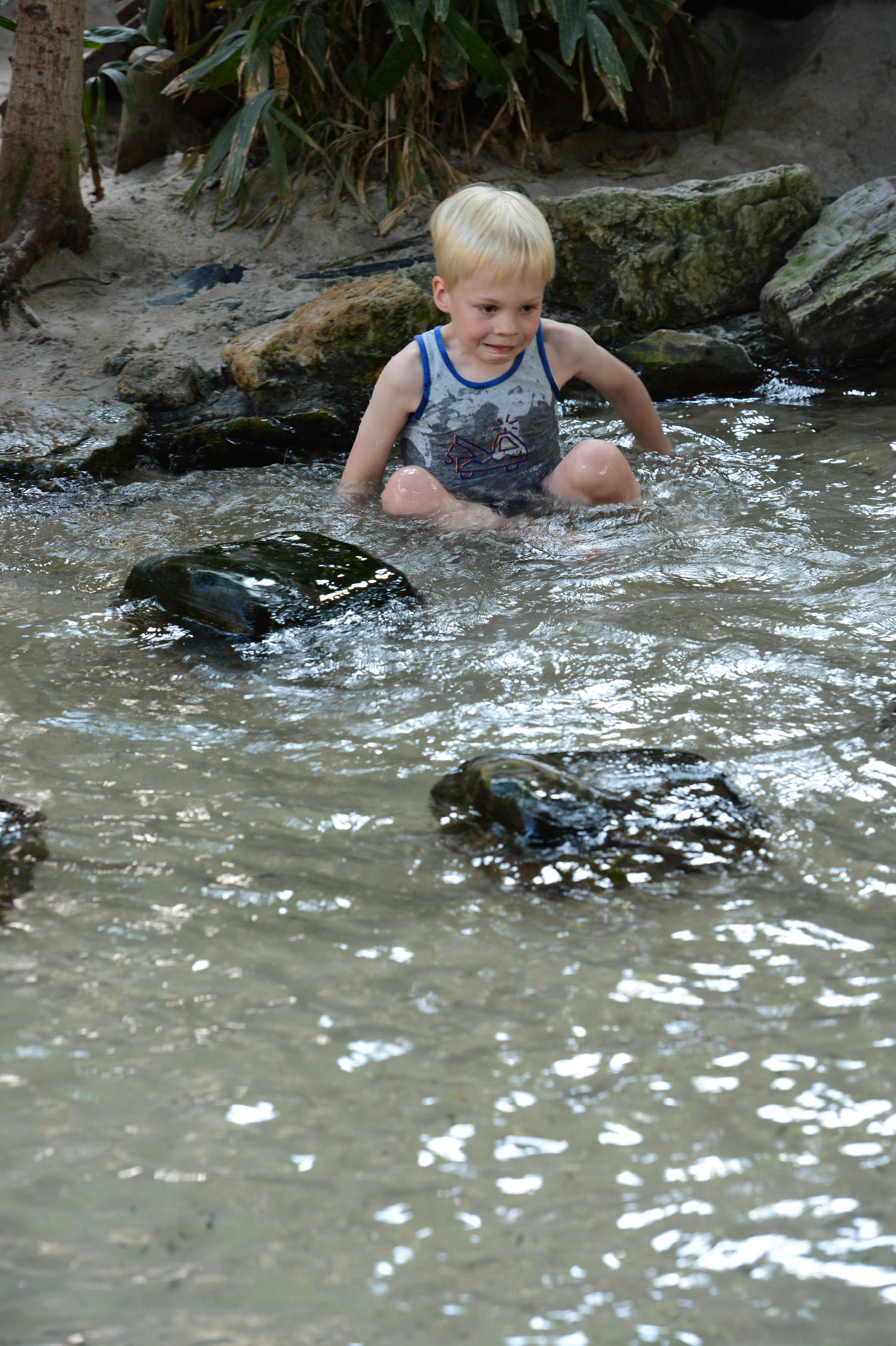 A young child sits in shallow water, surrounded by rocks, wearing a sleeveless shirt and looking focused.