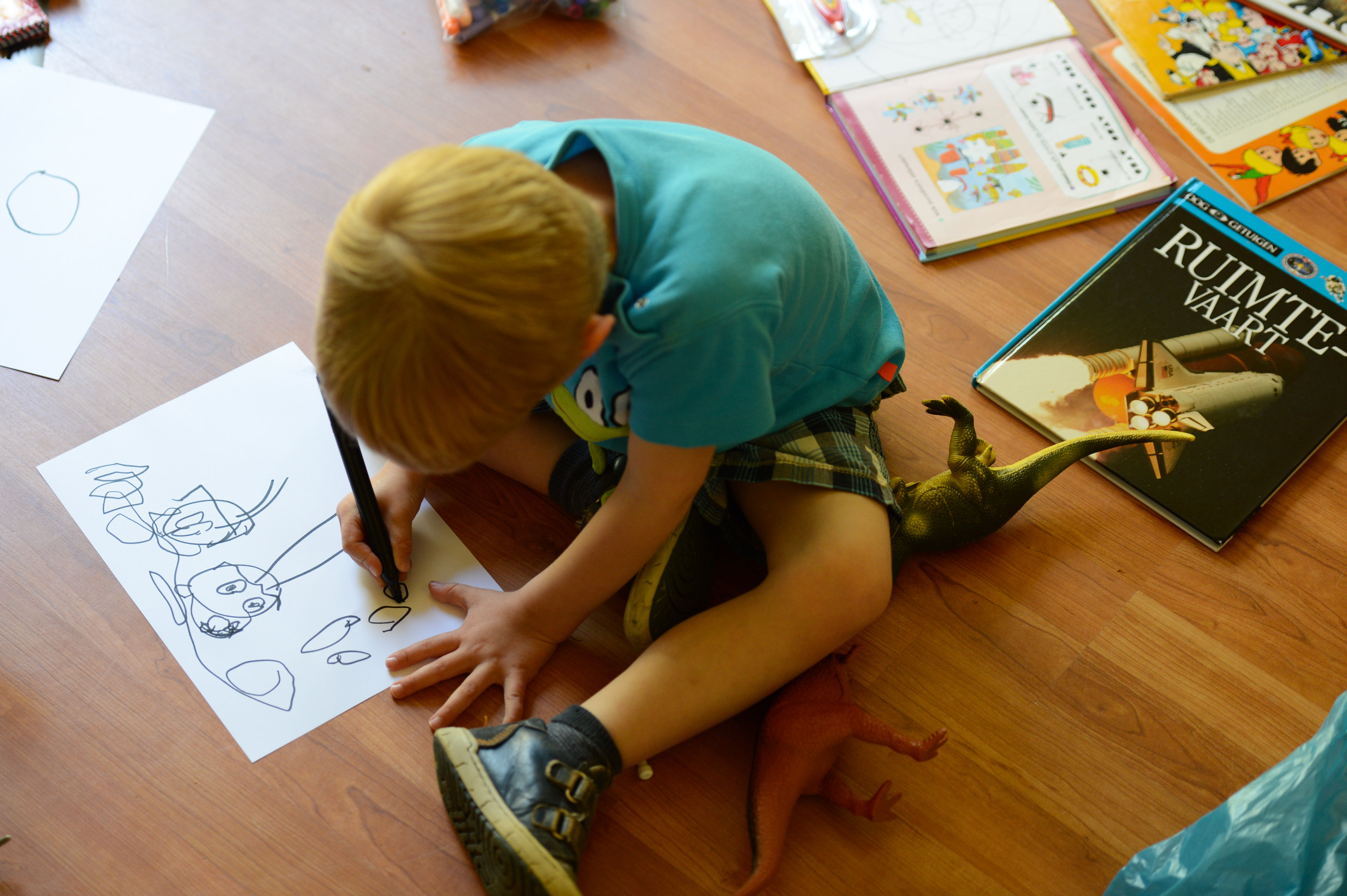 A young child sits on the floor, drawing with a black marker on paper, surrounded by books and toy dinosaurs.