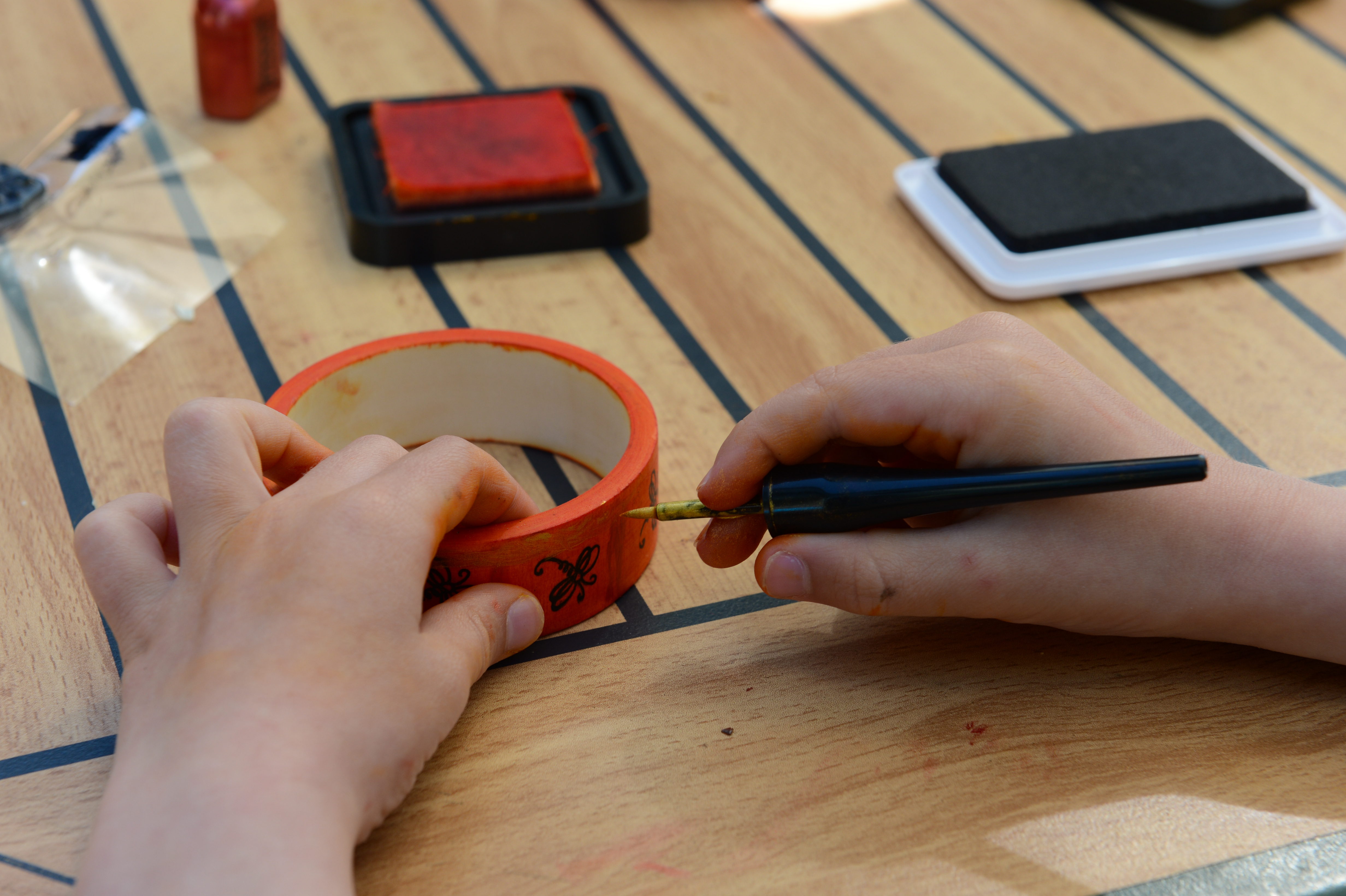 A child decorates a wooden bracelet with a black pen while sitting at a table with art supplies.