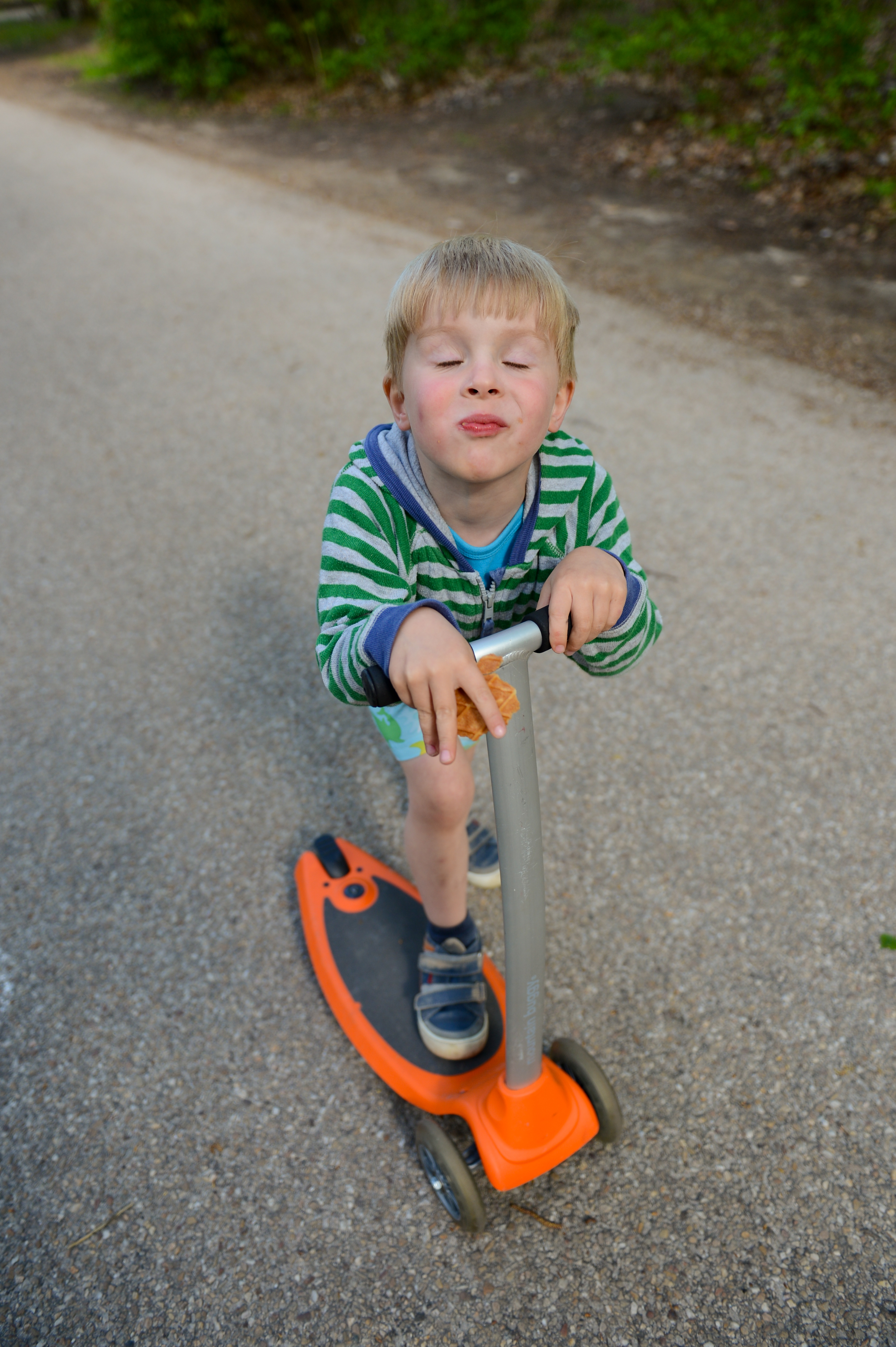 A young child in a striped hoodie rides an orange scooter on a paved path while holding a snack.