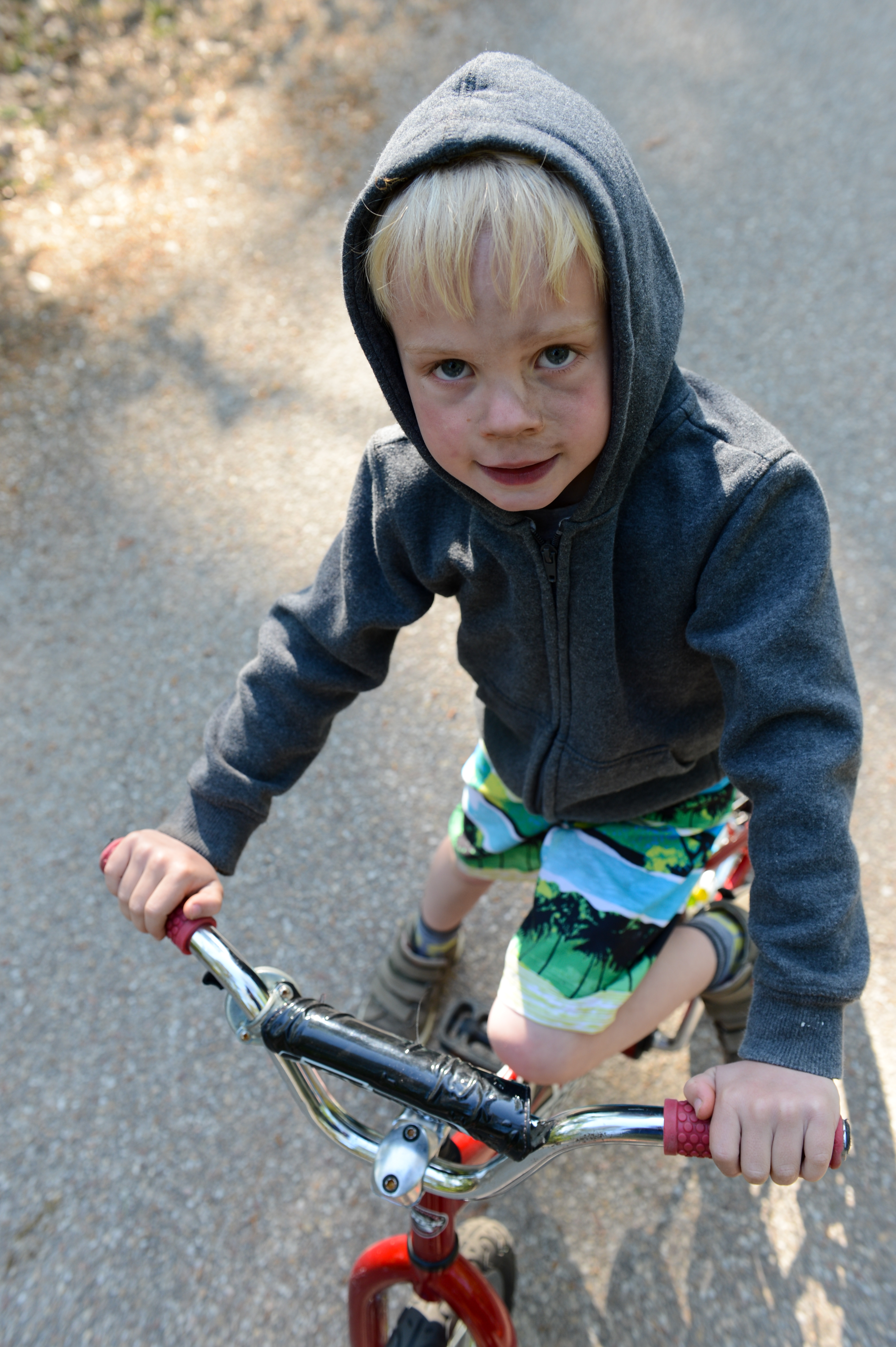 A young child in a hoodie rides a small red bike on a paved path, looking up at the camera.