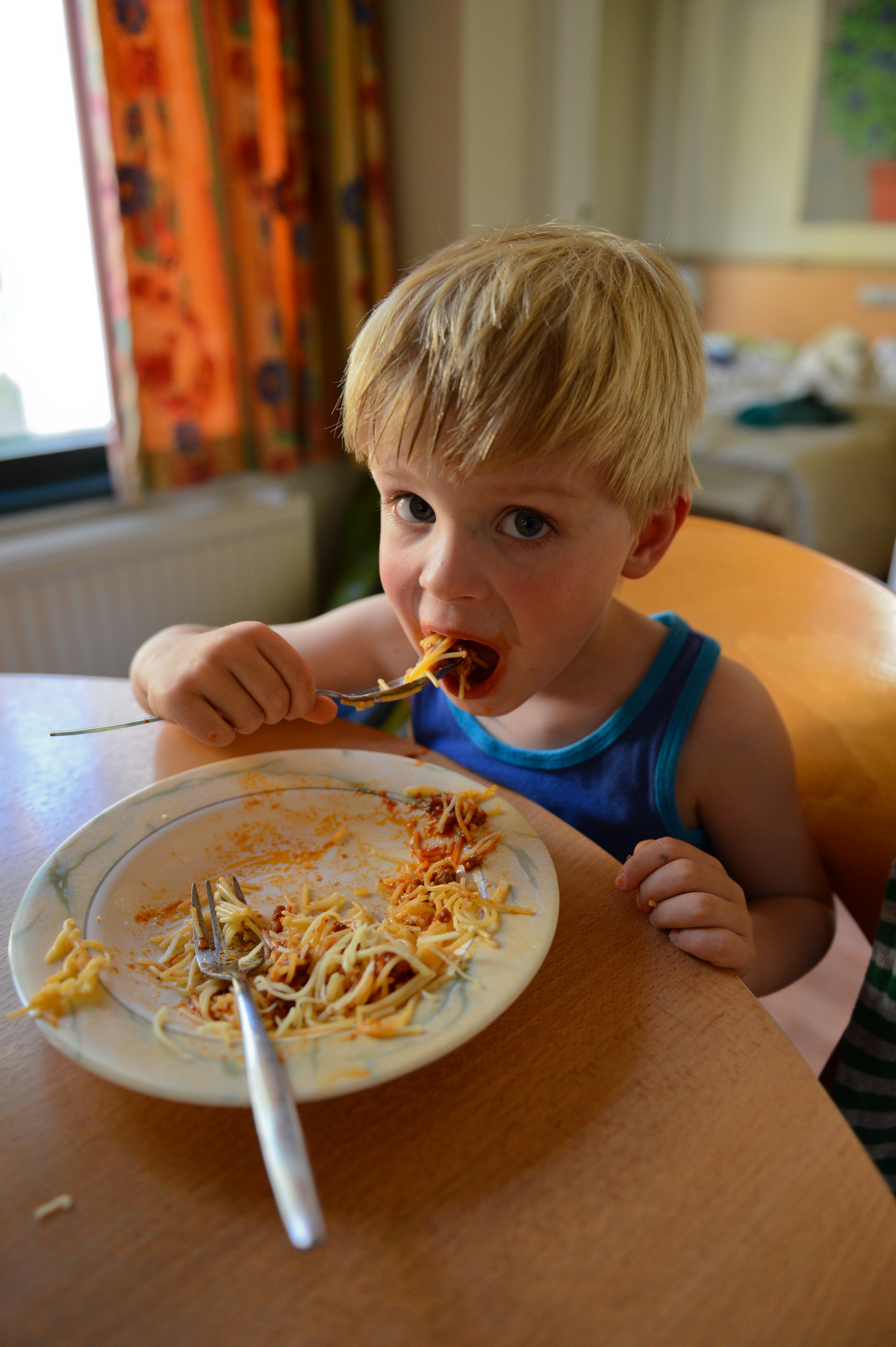 A young child in a blue tank top eats spaghetti at a table, using a fork.