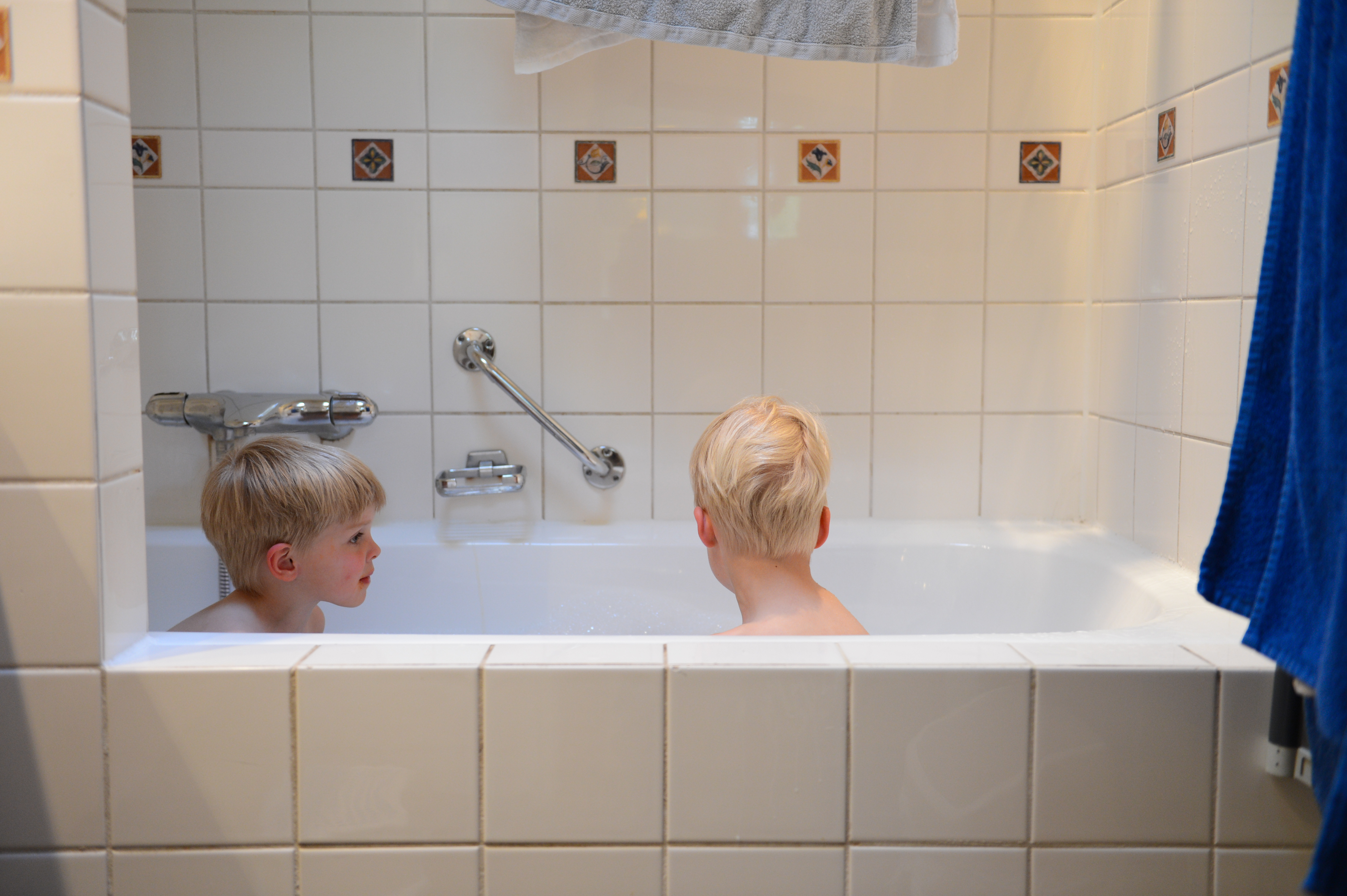 Two young children with light hair sit in a white bathtub, facing each other in a tiled bathroom.