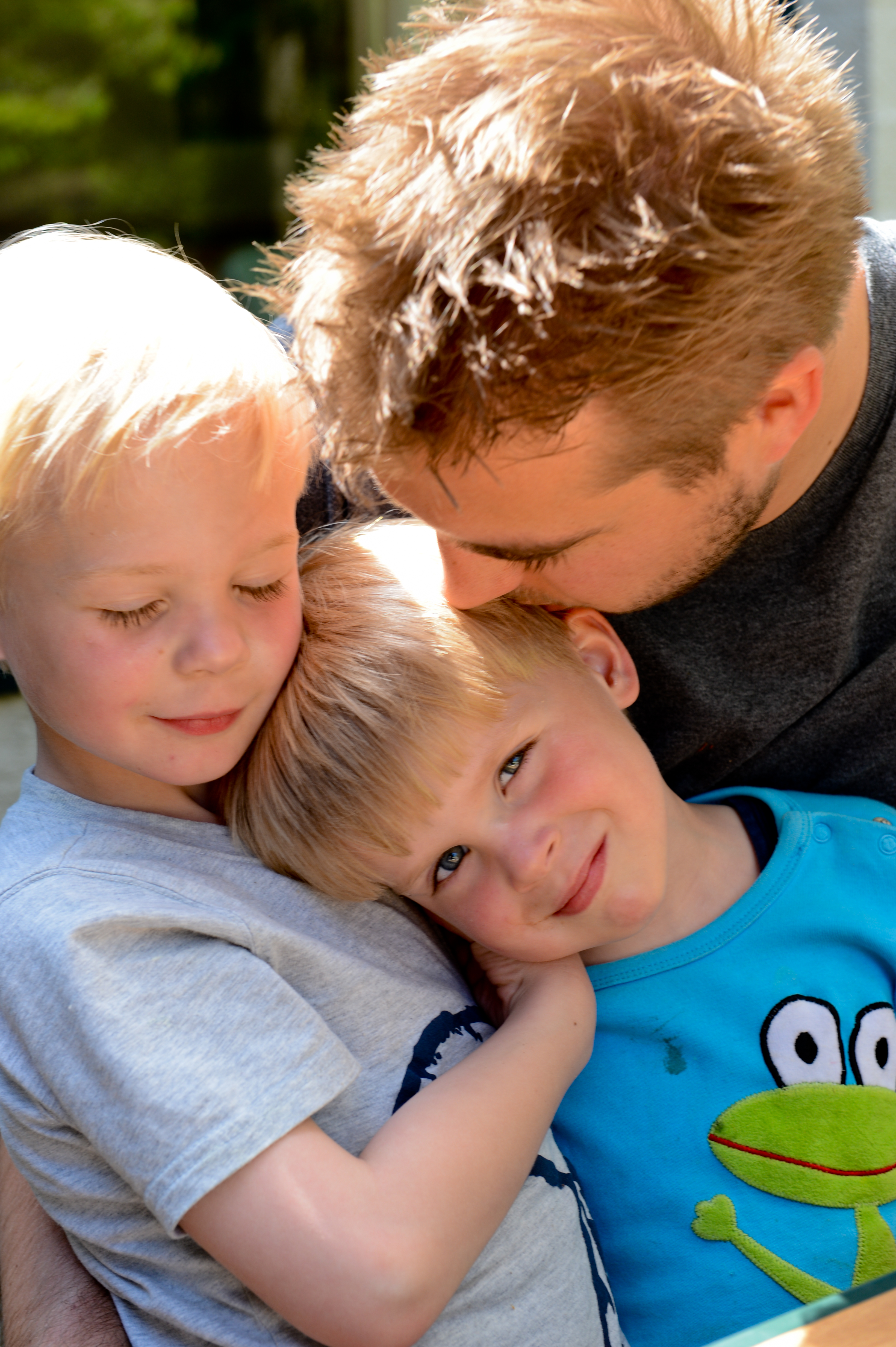A man kisses a young child's head while another child hugs them closely, all smiling and affectionate.