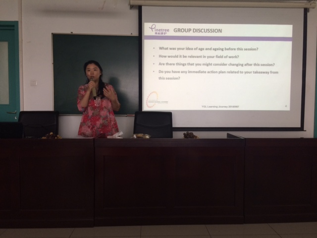 A woman stands at the front of a classroom, speaking into a microphone during a group discussion.