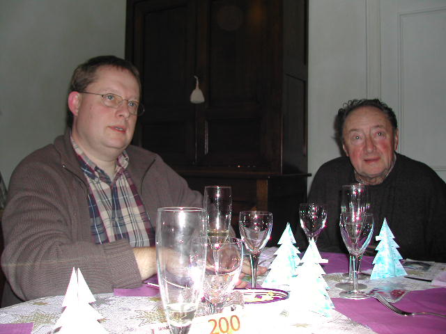 Two men sitting at a table with glasses and small paper Christmas trees, engaged in conversation.